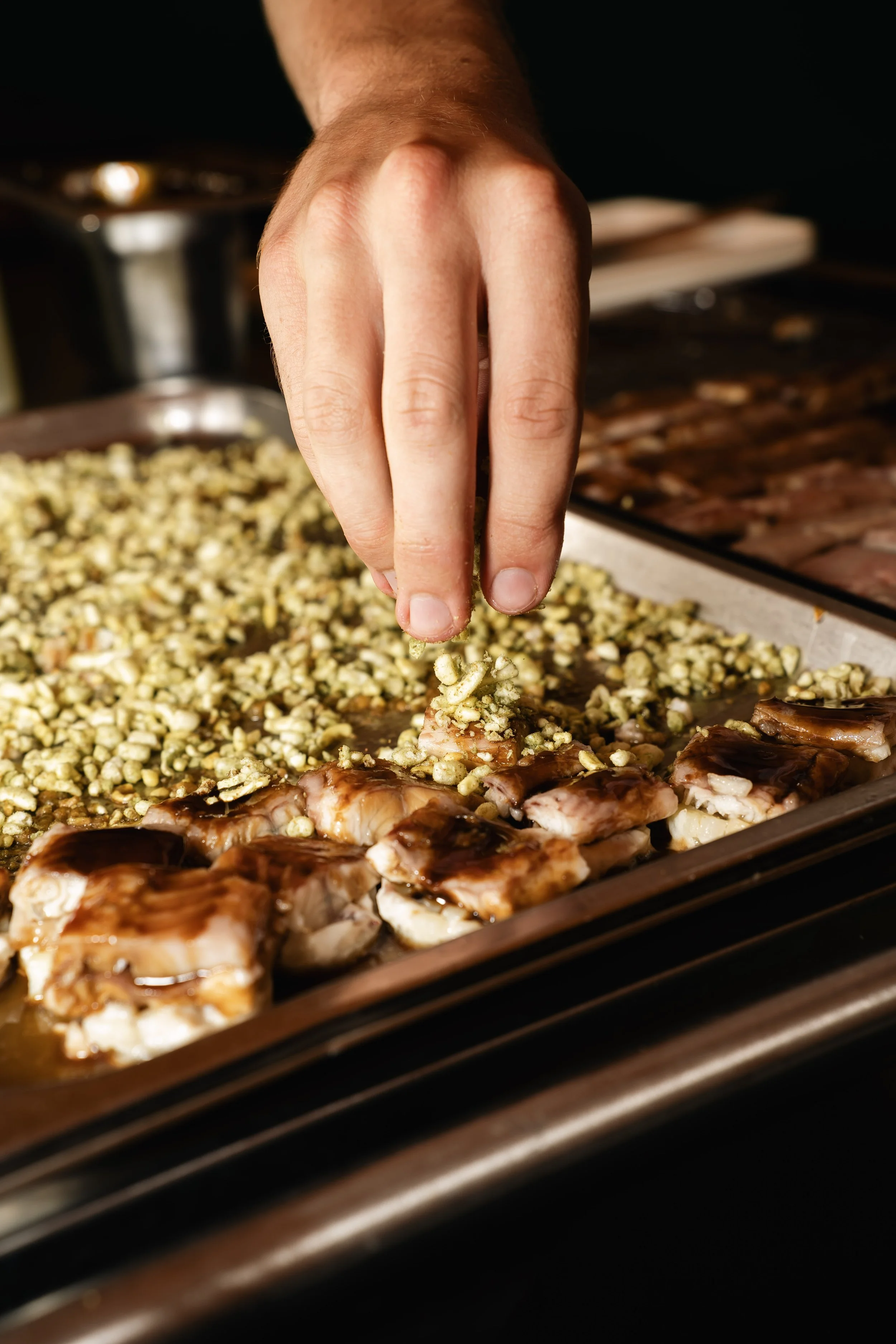Een hand strooit kruiden over gegrilde kippenvleugels en rijst met groenten op een bakplaat.