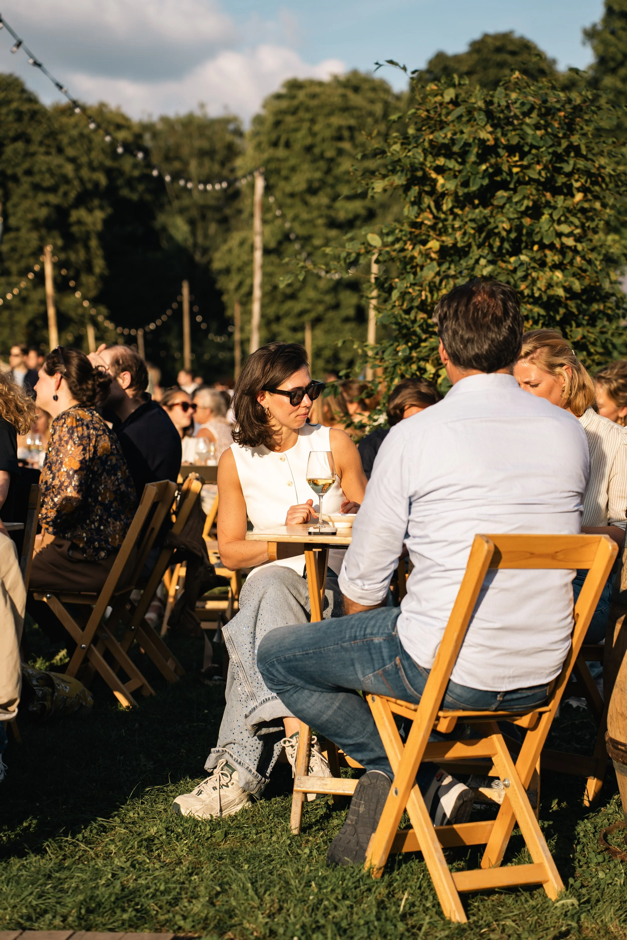 Mensen zitten buiten op houten stoelen in een groene tuin tijdens een feestje, genieten van de zon met een glas witte wijn.