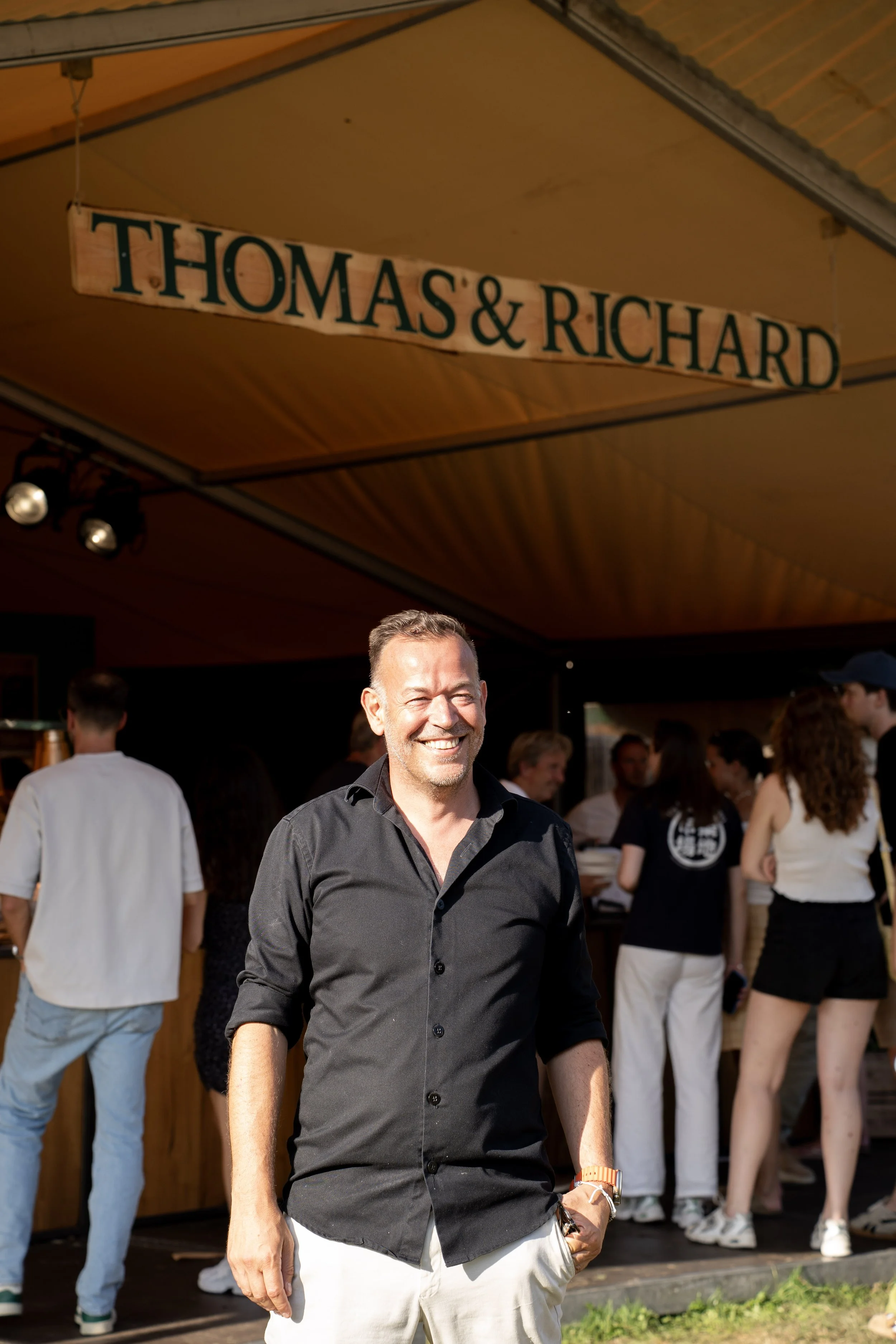 Man in zwart overhemd en witte broek lachend op een feestje met mensen op de achtergrond