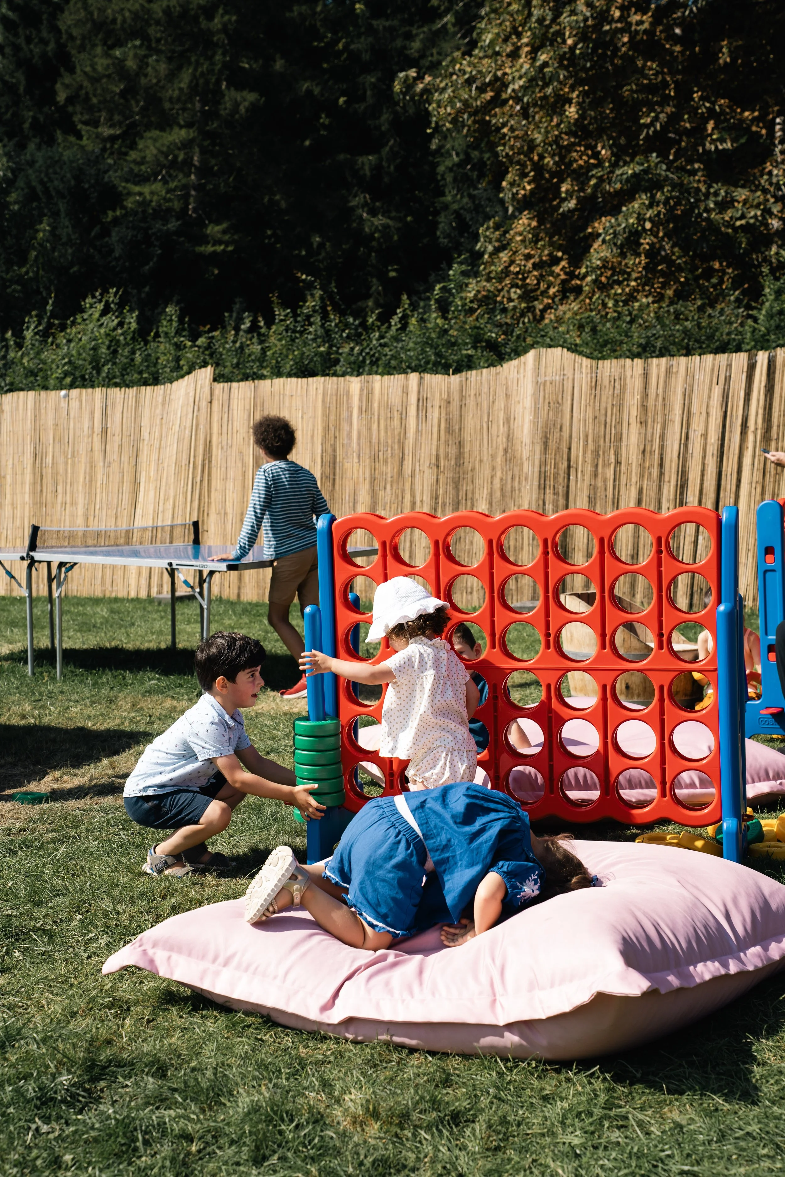 Kinderen spelen buiten in de tuin, met een groot rood-oranje spelhuis en een schommel, terwijl een ander kind gebruik maakt van een tafeltennistafel.