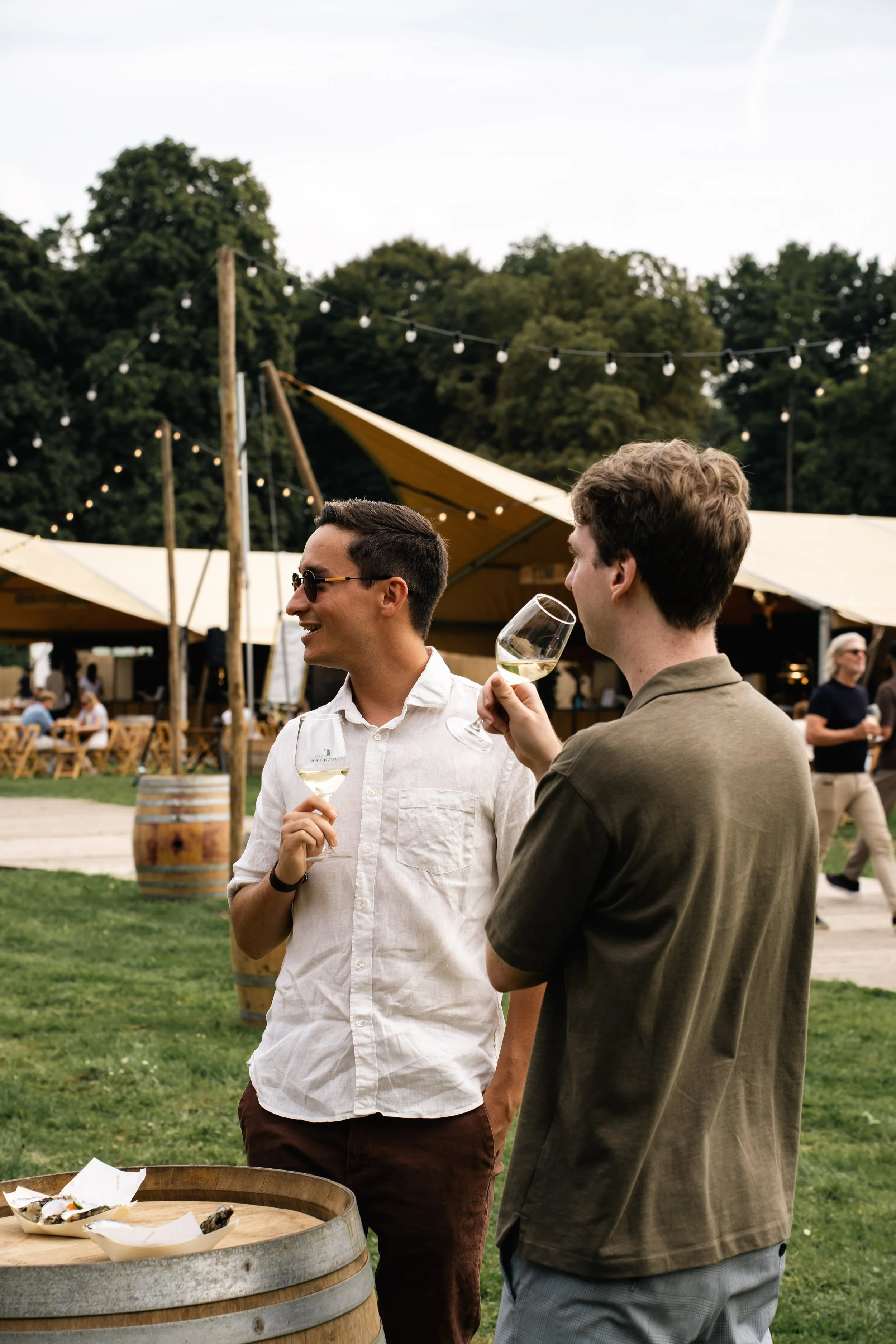 Twee mannen genieten van een drankje op een buitenevenement met tenten en bollen in de lucht, omgeven door groen en bomen.