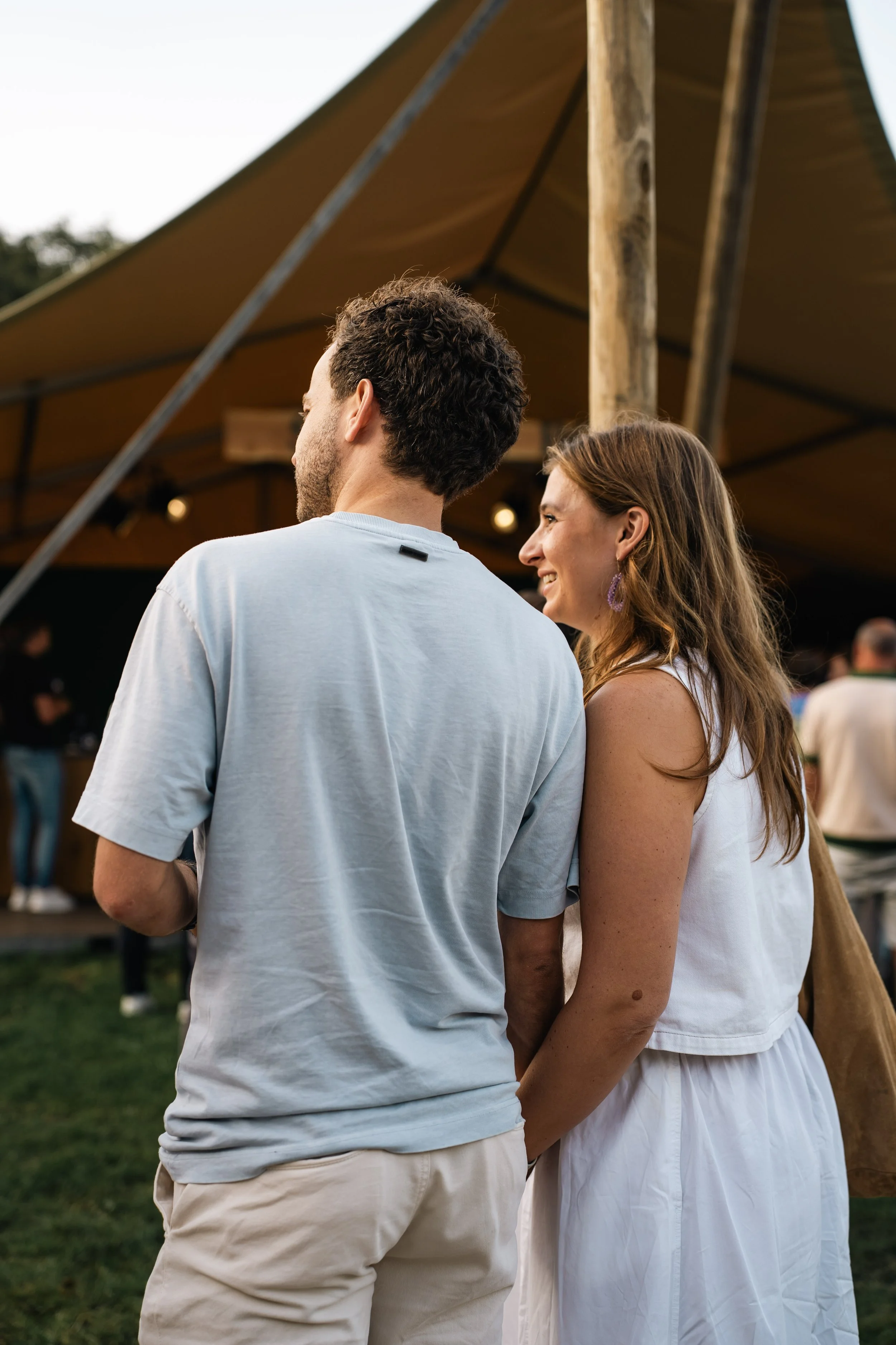 Twee mensen staan bij elkaar in een buitenruimte met een tent en andere mensen op de achtergrond tijdens een evenement.