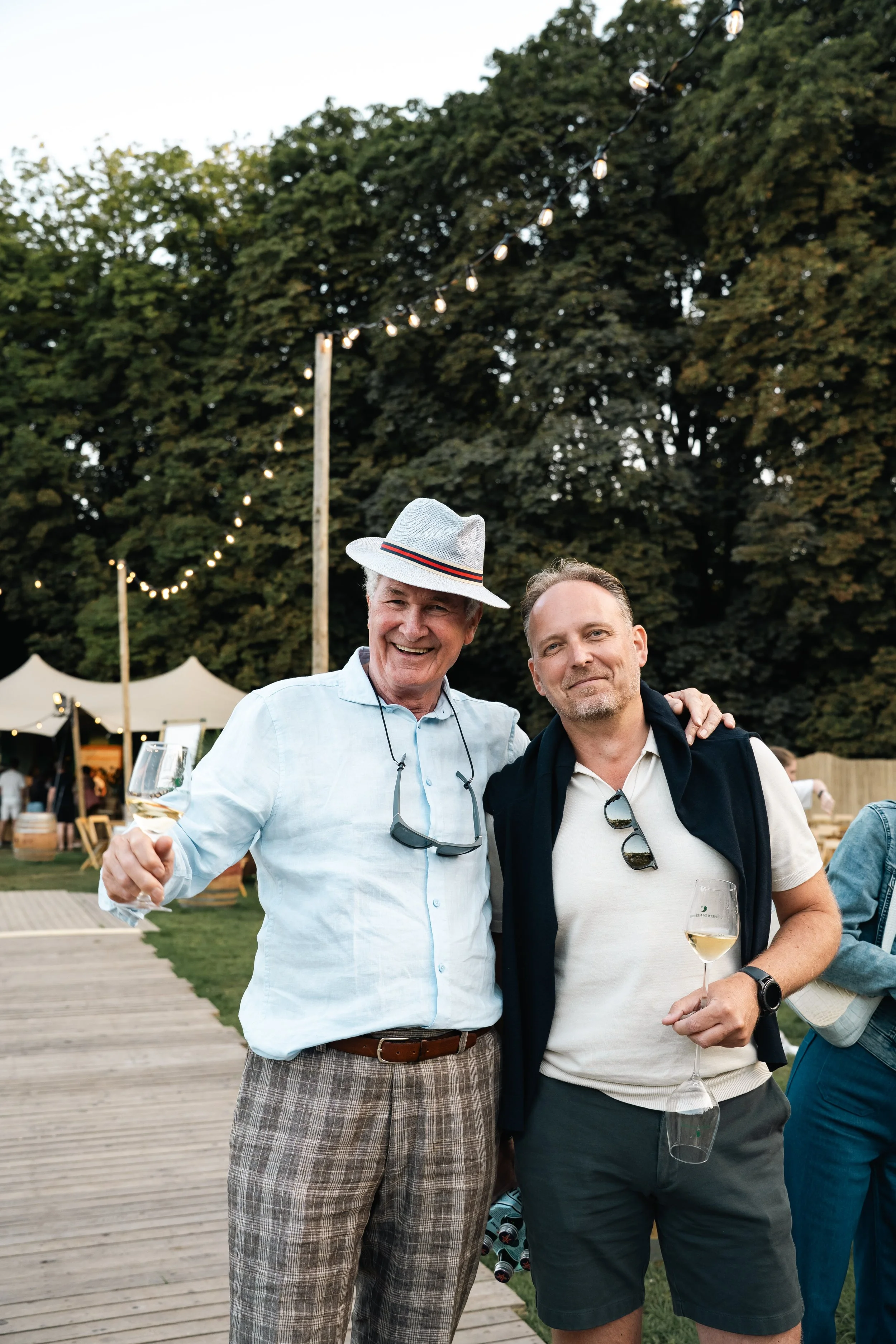 Twee mannen die samen genieten op een buitenfeest, met één die een glas witte wijn vasthoudt, onder een hemel met sfeervolle verlichting en groene bomen op de achtergrond.