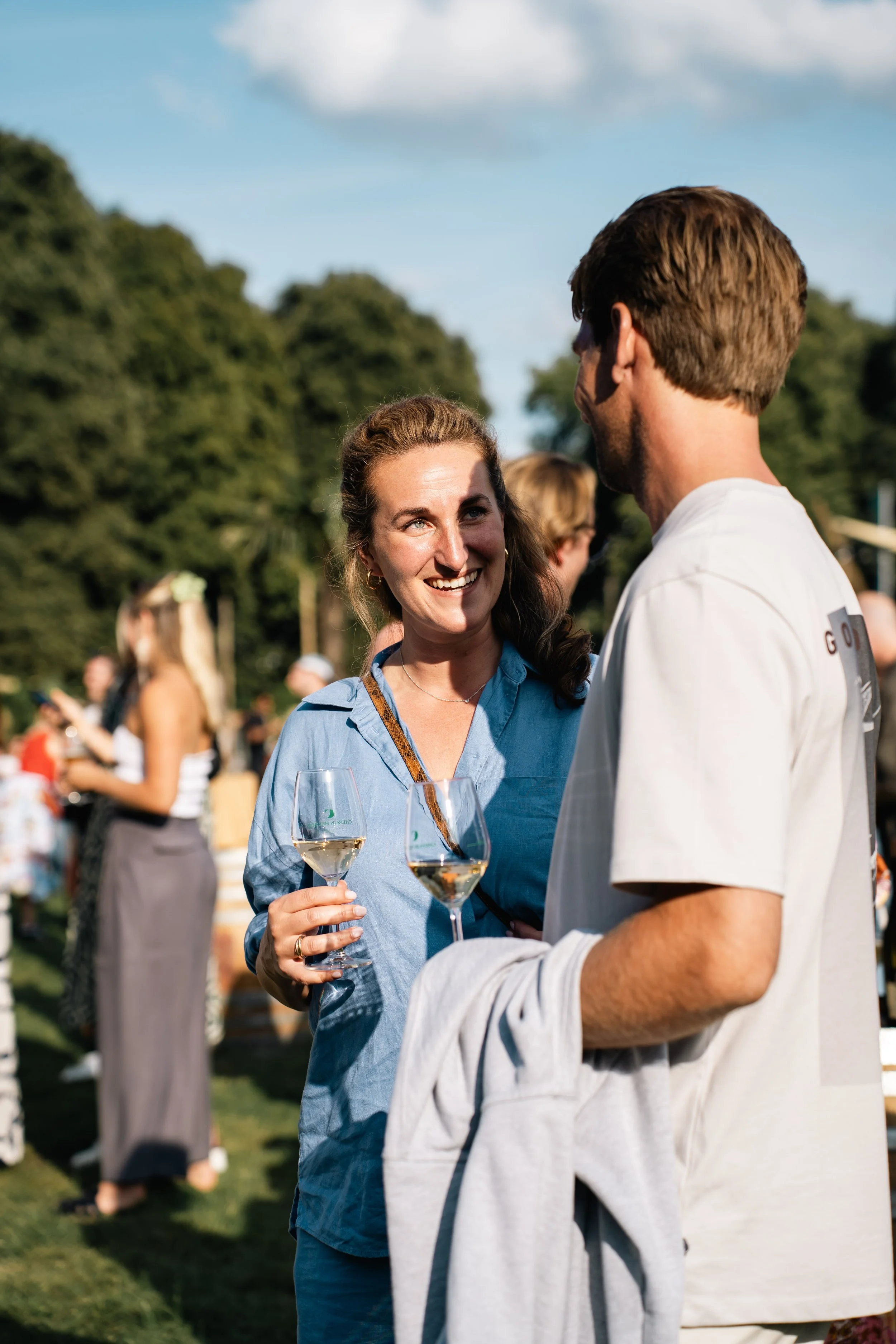 Twee mensen die in de buitenlucht wijn drinken en praten tijdens een feest of bijeenkomst, met andere mensen op de achtergrond.