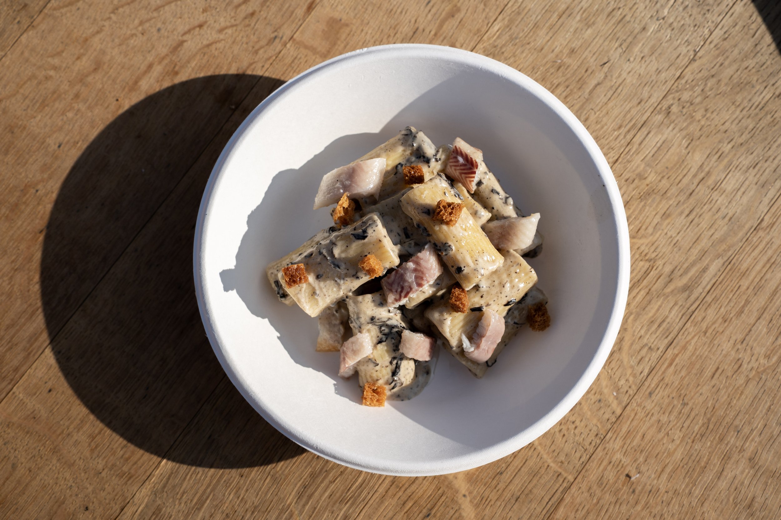 Een wit bord met pasta salade en stukjes kipmuton, bestrooid met kleine krokante croutons, op een houten tafel in de zon.