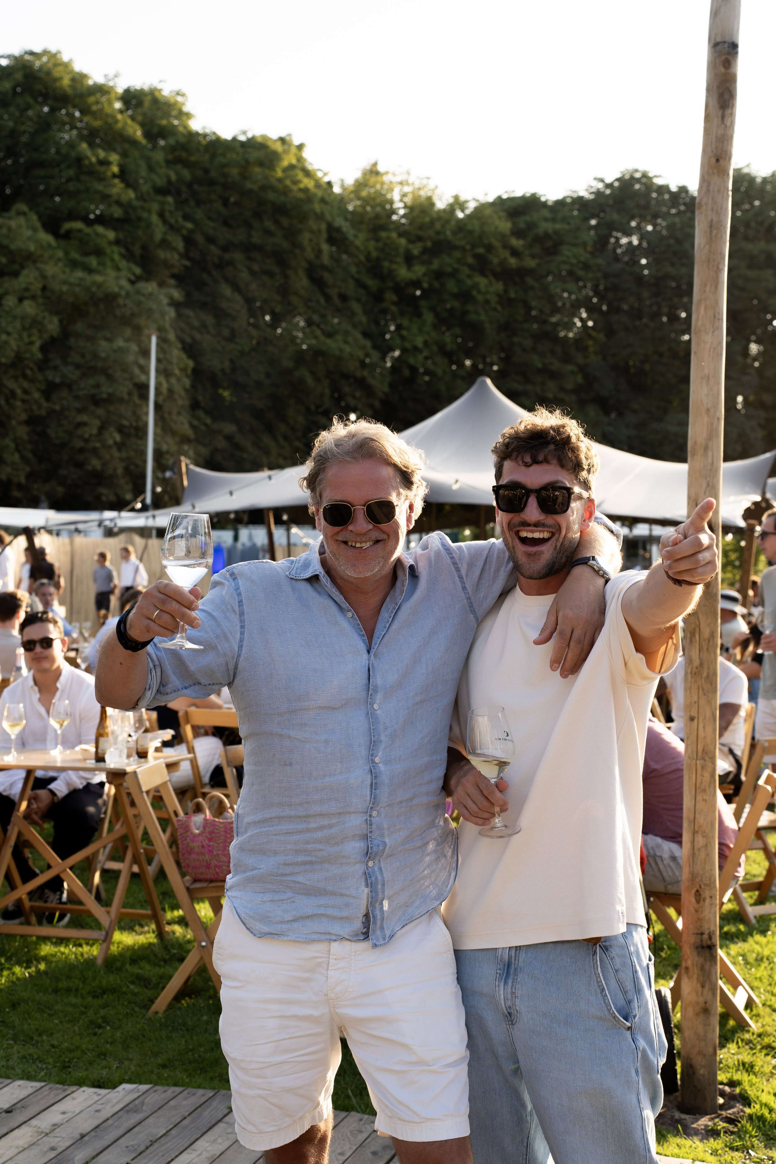 Twee mannen glimlachen, dragen zonnebrillen, en houden glazen witte wijn vast tijdens een buitenfeest met witte tenten en mensen op de achtergrond.