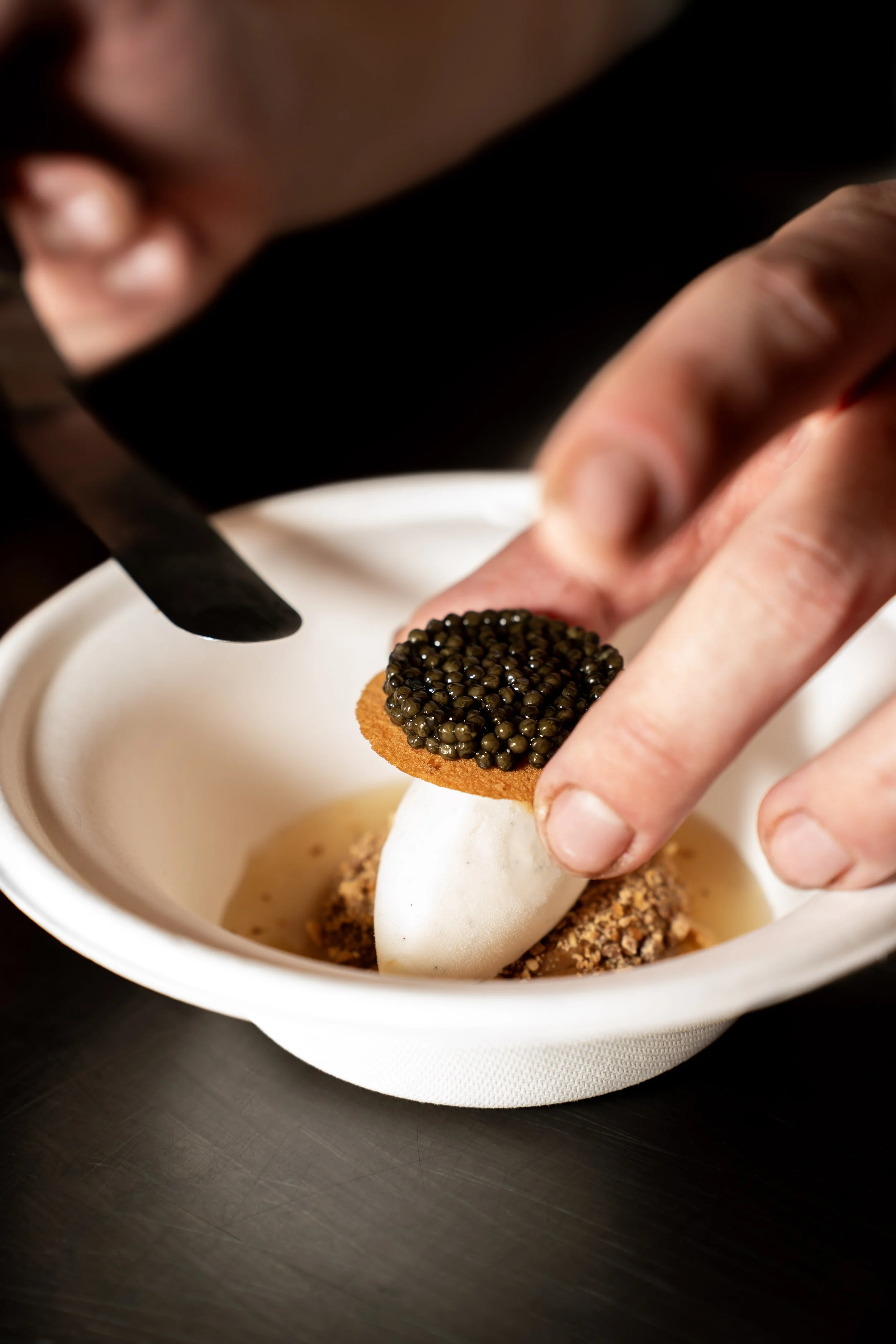 Een hand plaatst een kaviaar beleg op een wit aperitiefbuisje dat in een kom met een saus zit.