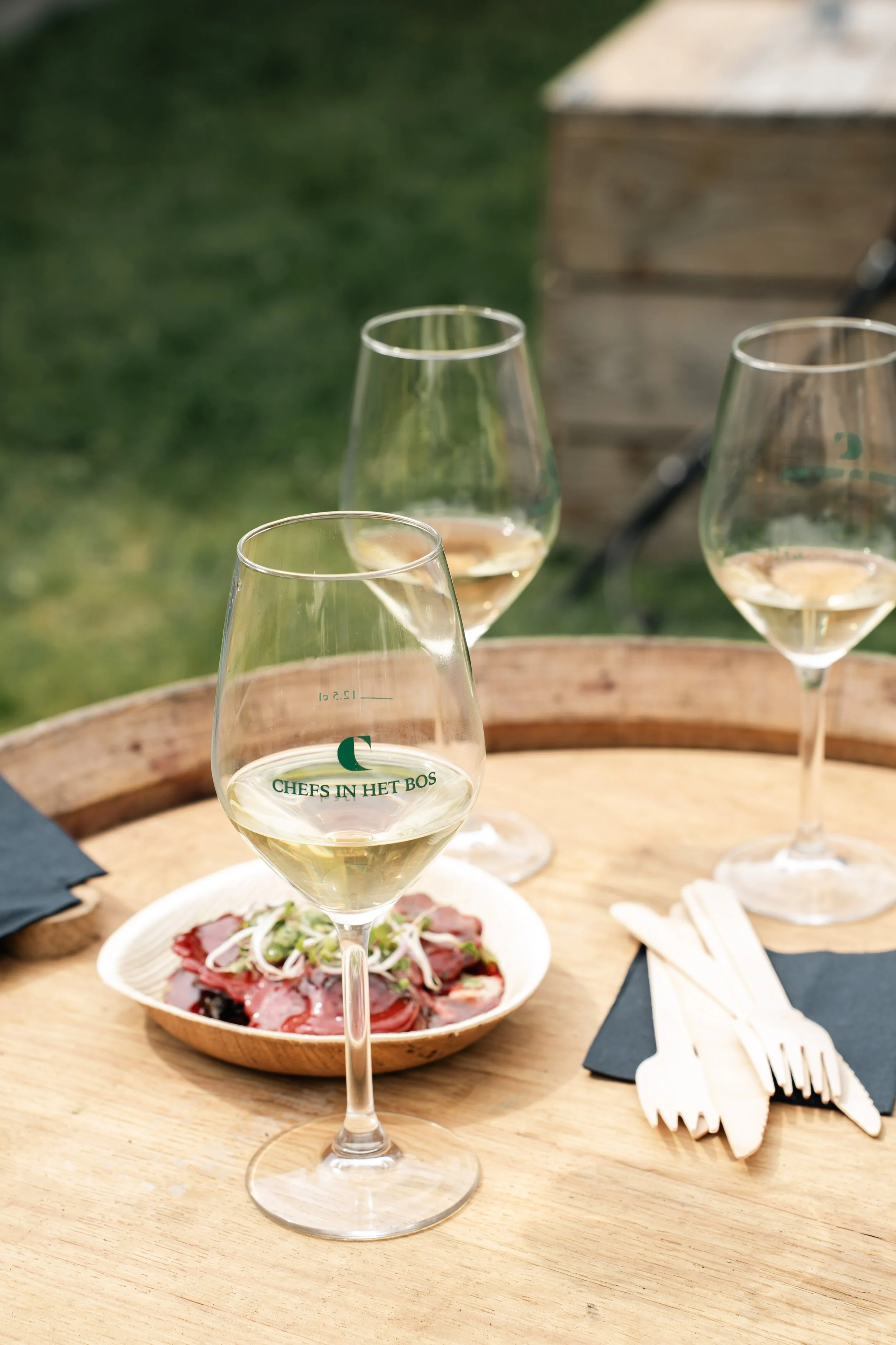 Drie wijnglazen met witte wijn op een houten tafel, met een bord met vlees en groenten, servetten, vorken en messen, in een buitenomgeving.