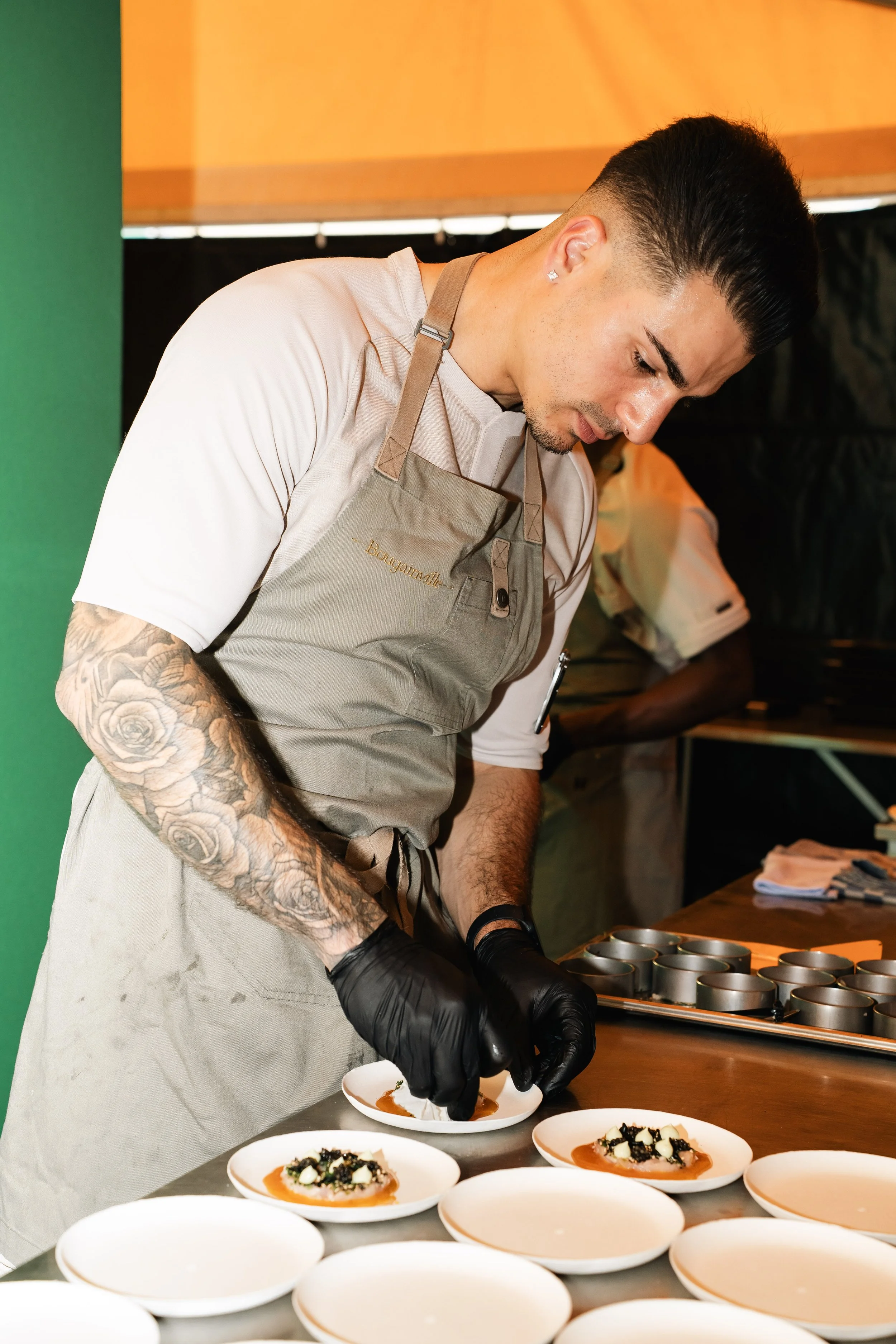 Een chef-kok in een beige uniform en zwarte handschoenen versiert borden met gerecht in een keuken, met meerdere lege witte borden op de tafel voor hem.
