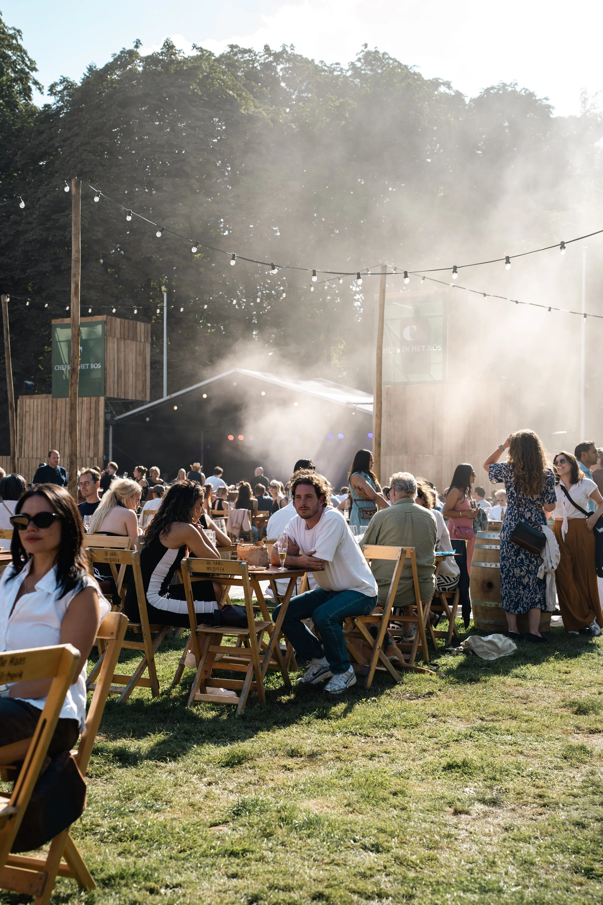 Mensen genieten van een buitenfestival op een grasveld met houten stoelen, in de achtergrond bomen en een houten structuur, met een lichte mist en zonlicht