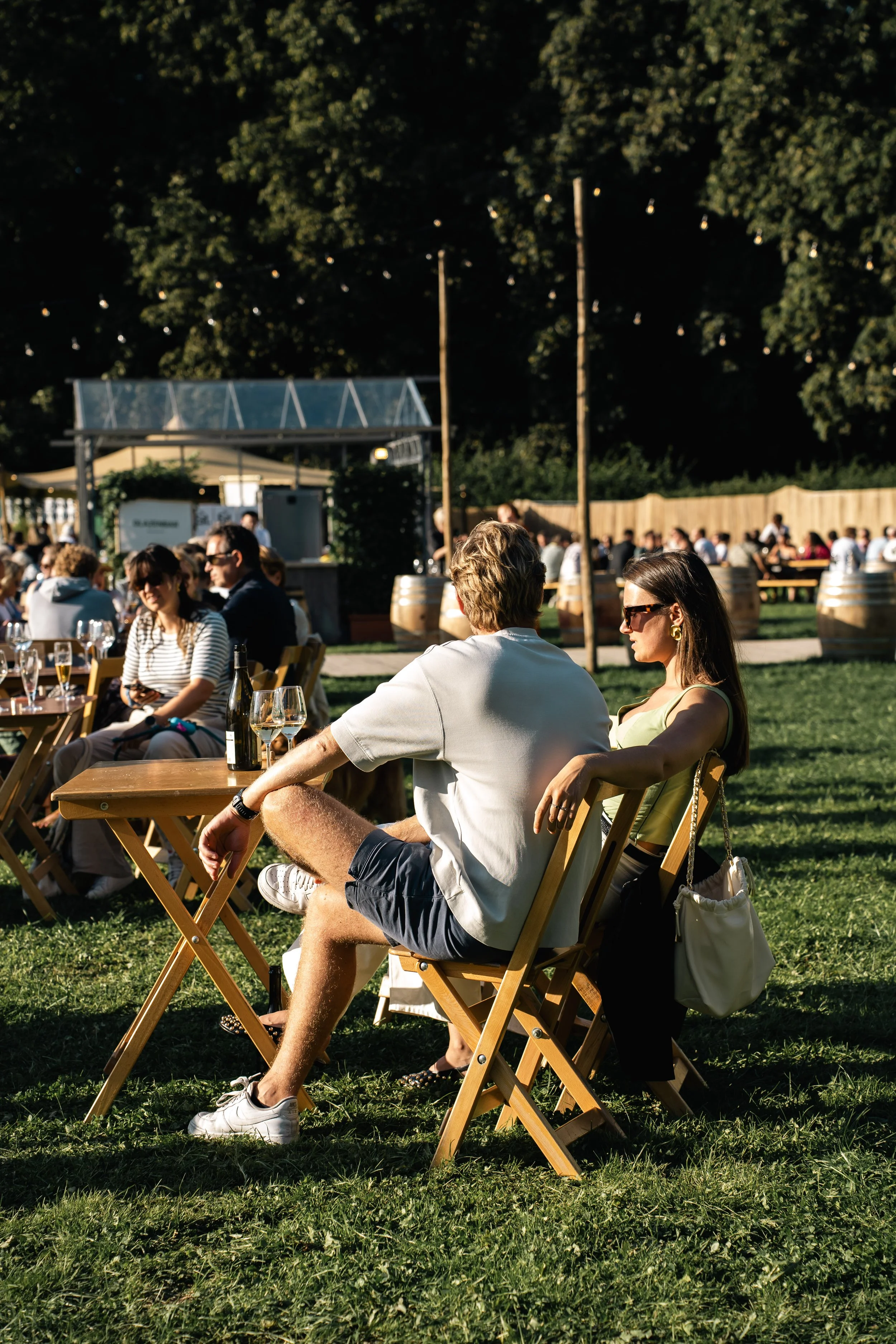 Mensen zitten buiten op een terras op een zonnige dag, sommigen dragen zonnebrillen en genieten van drankjes, op de achtergrond staan grote wijnvaten en boomverlichting.