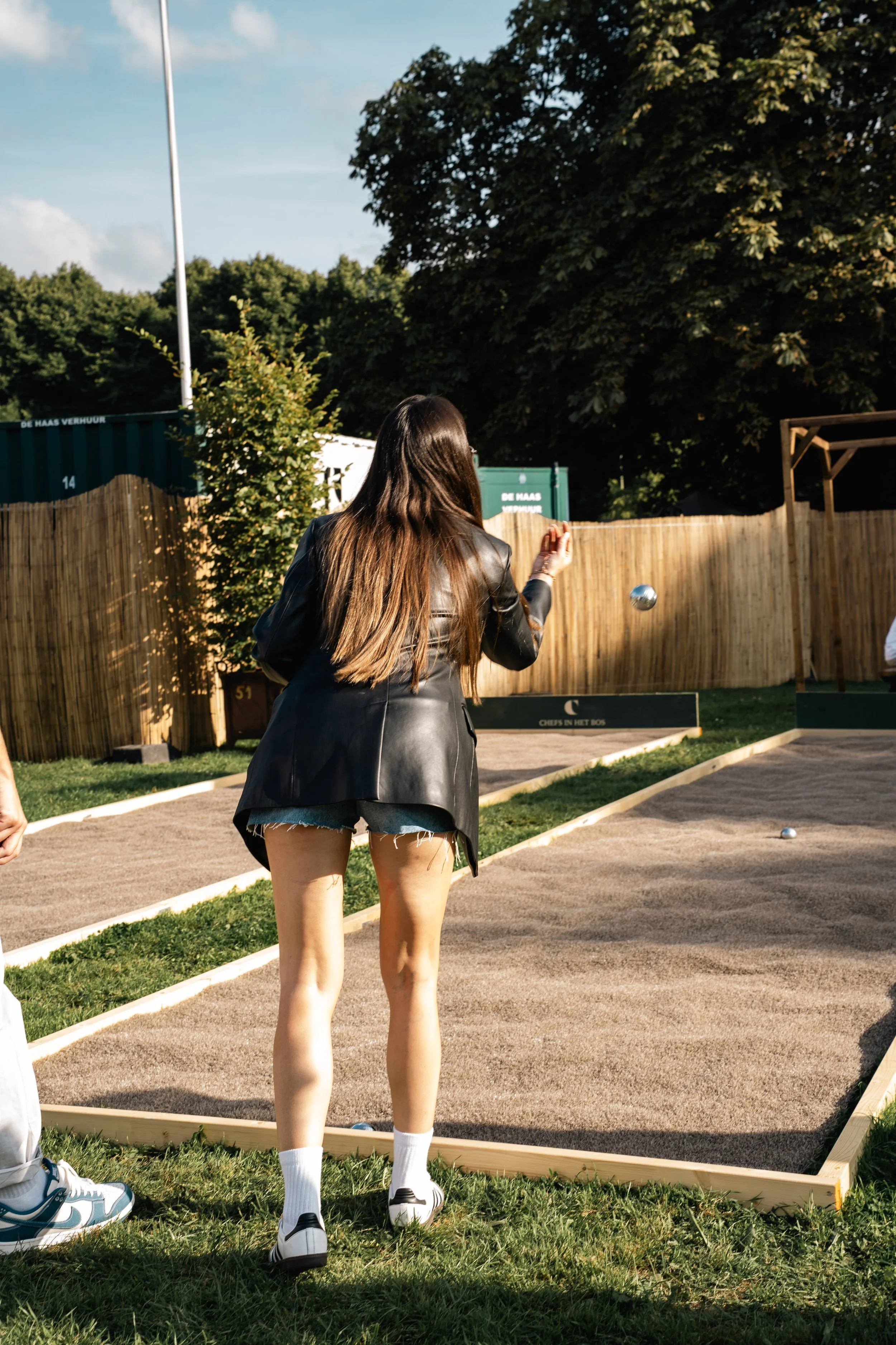 Een vrouw met lang donker haar in een zwart leren jacket en korte denim shorts, die een ijzeren bal gooit op een petanquebaan in een tuin, omringd door een houten hek en groene bomen onder een blauwe hemel.