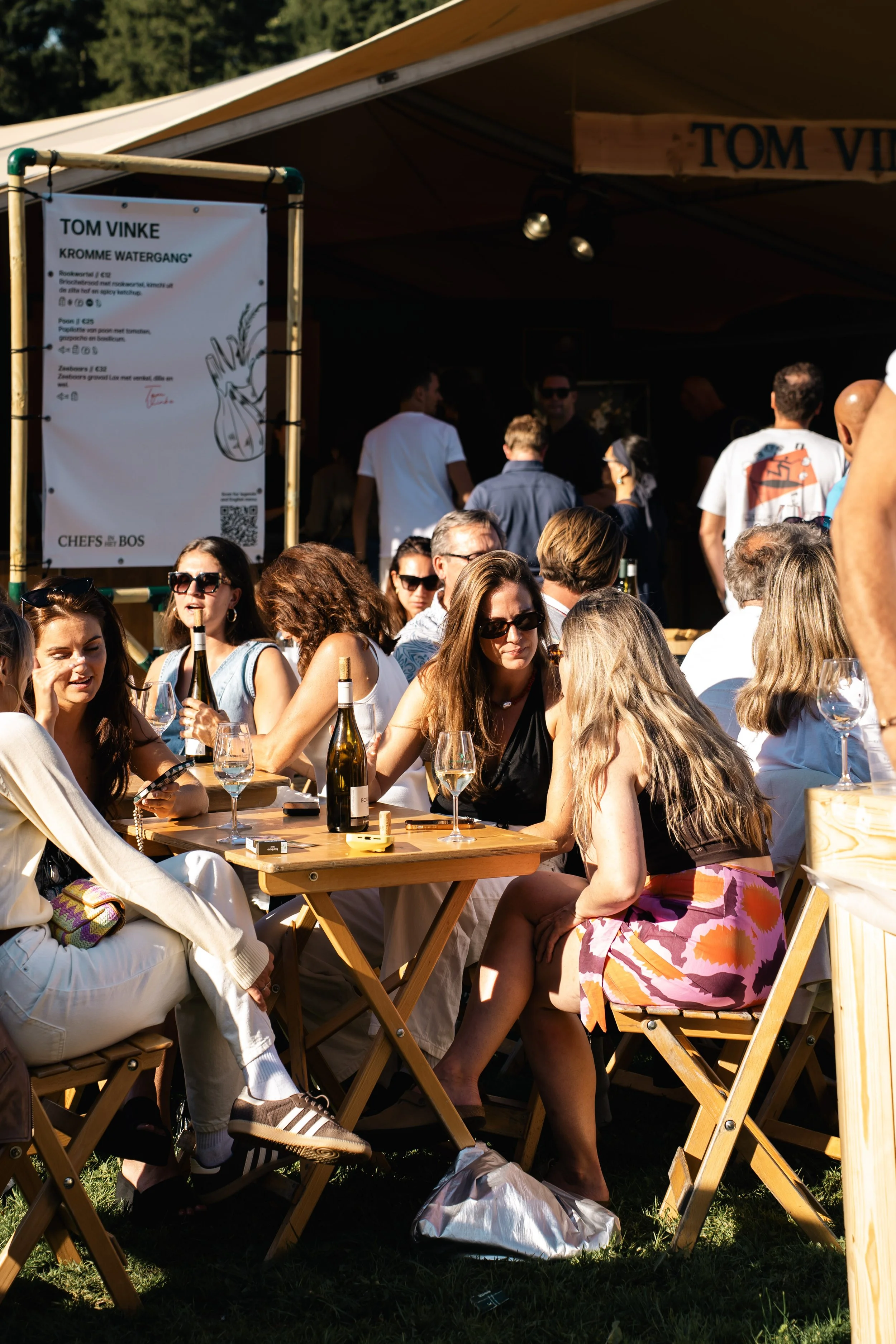 Een groep mensen zit buiten op houten stoelen aan een tafel, met glazen witte wijn en eten, in de zon. Achter hen is een terras met een grote menubord en een houten overkapping.