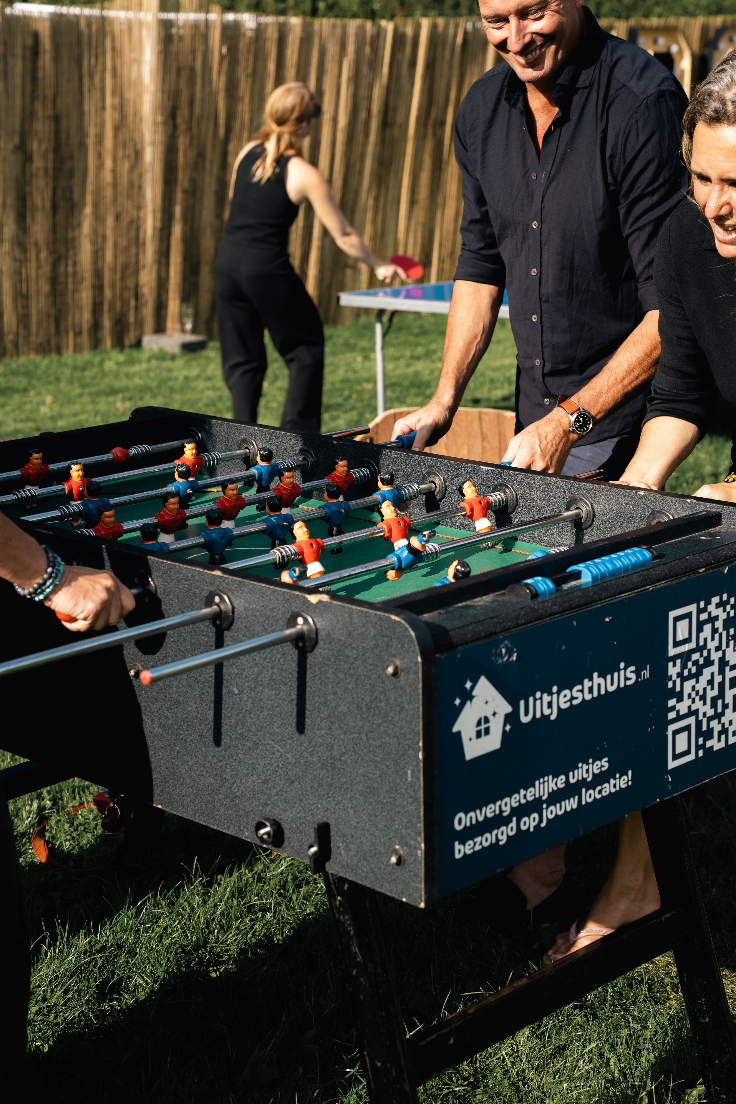 Een groep mensen die samen tafelvoetbal spelen in een achtertuin op gras, met een houten schutting op de achtergrond.