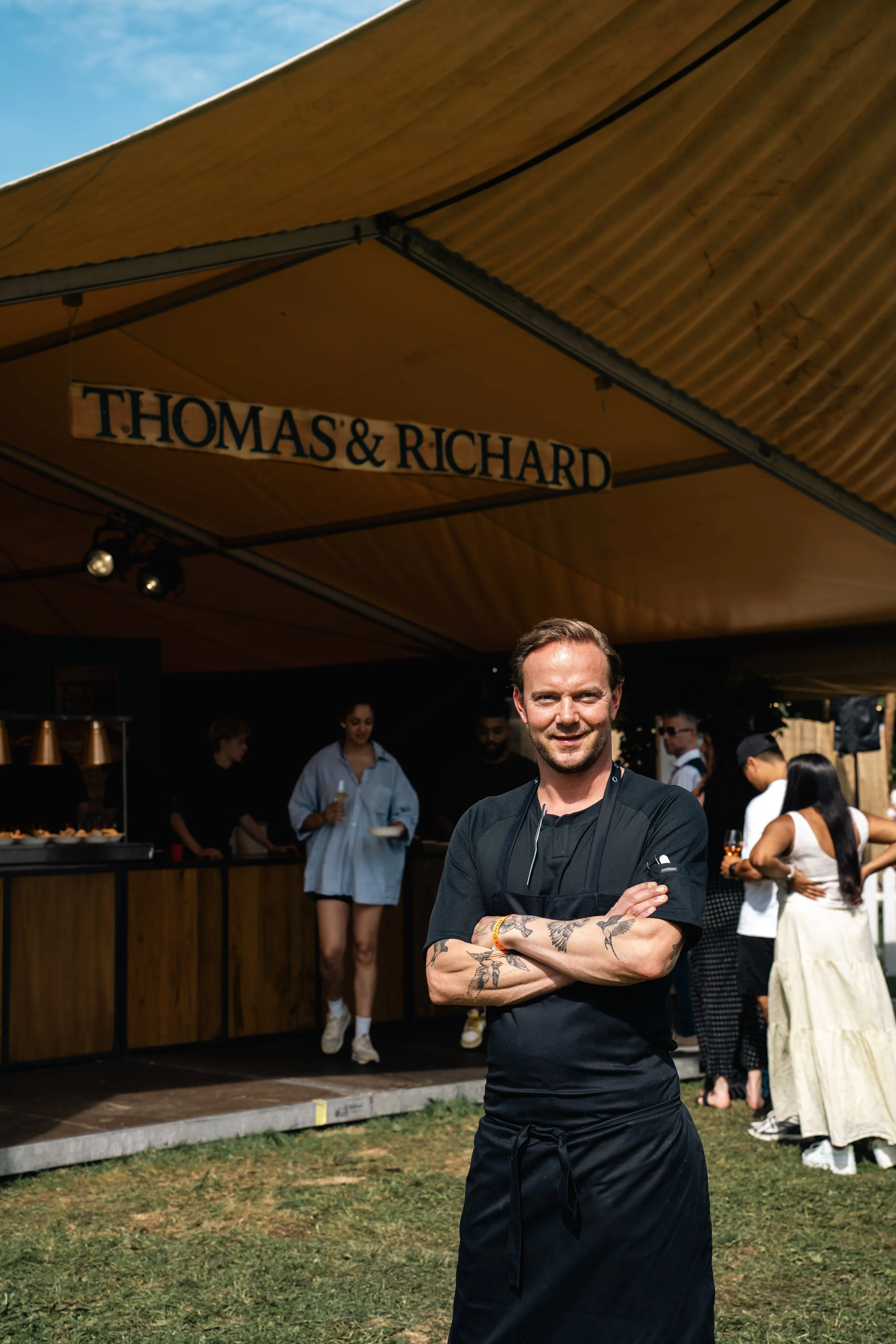 Man met tatoeages op armen, gekleed in zwart uniform, staat met gekruiste armen voor een foodtruck met de naam Thomas & Richard, mensen wachten op eten op een zonnig evenement.