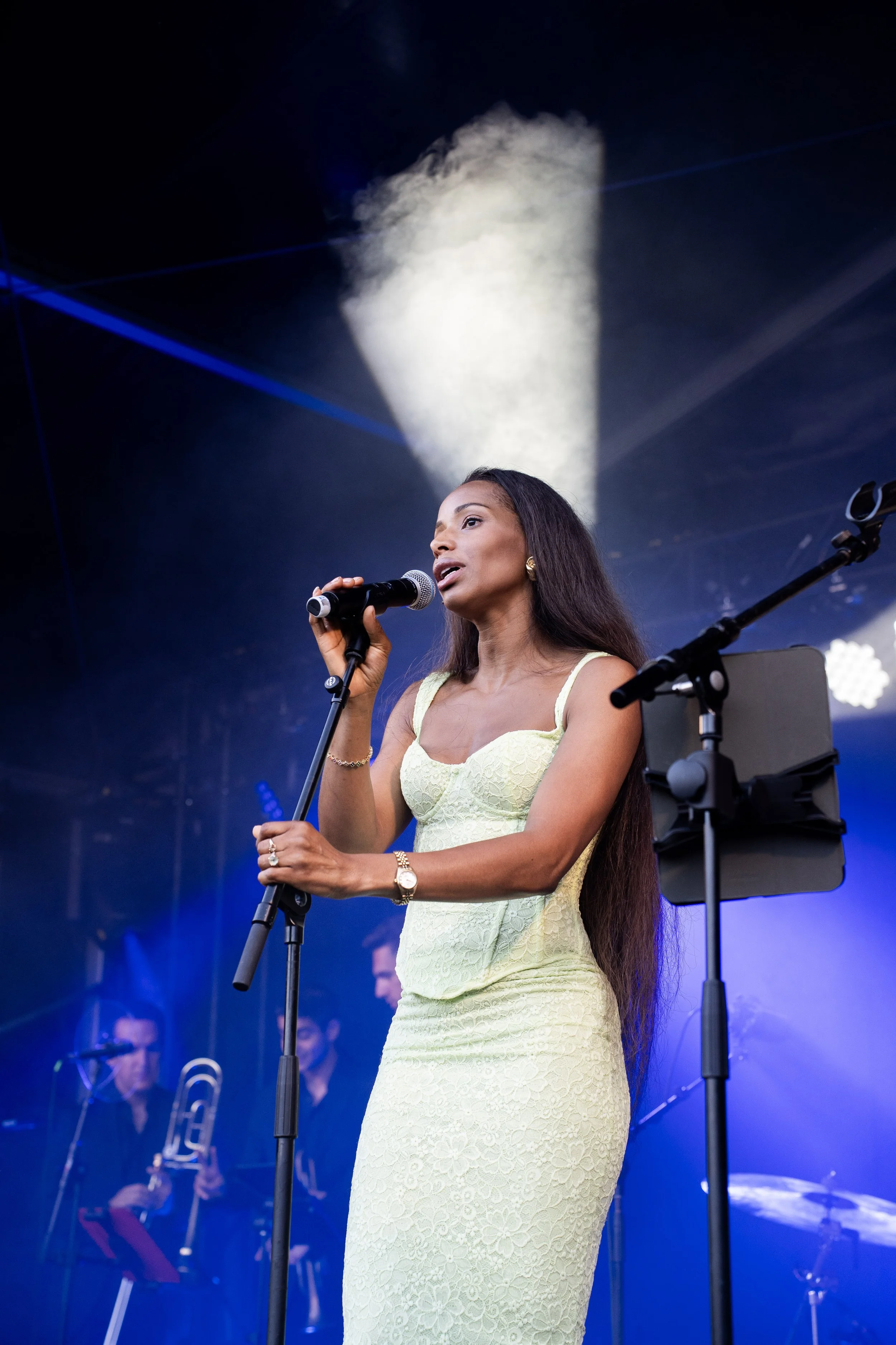 Een vrouw zingt in een geel kanten jurk tijdens een podiumoptreden met muzikanten op de achtergrond, verlicht door blauwe en witte lichten.