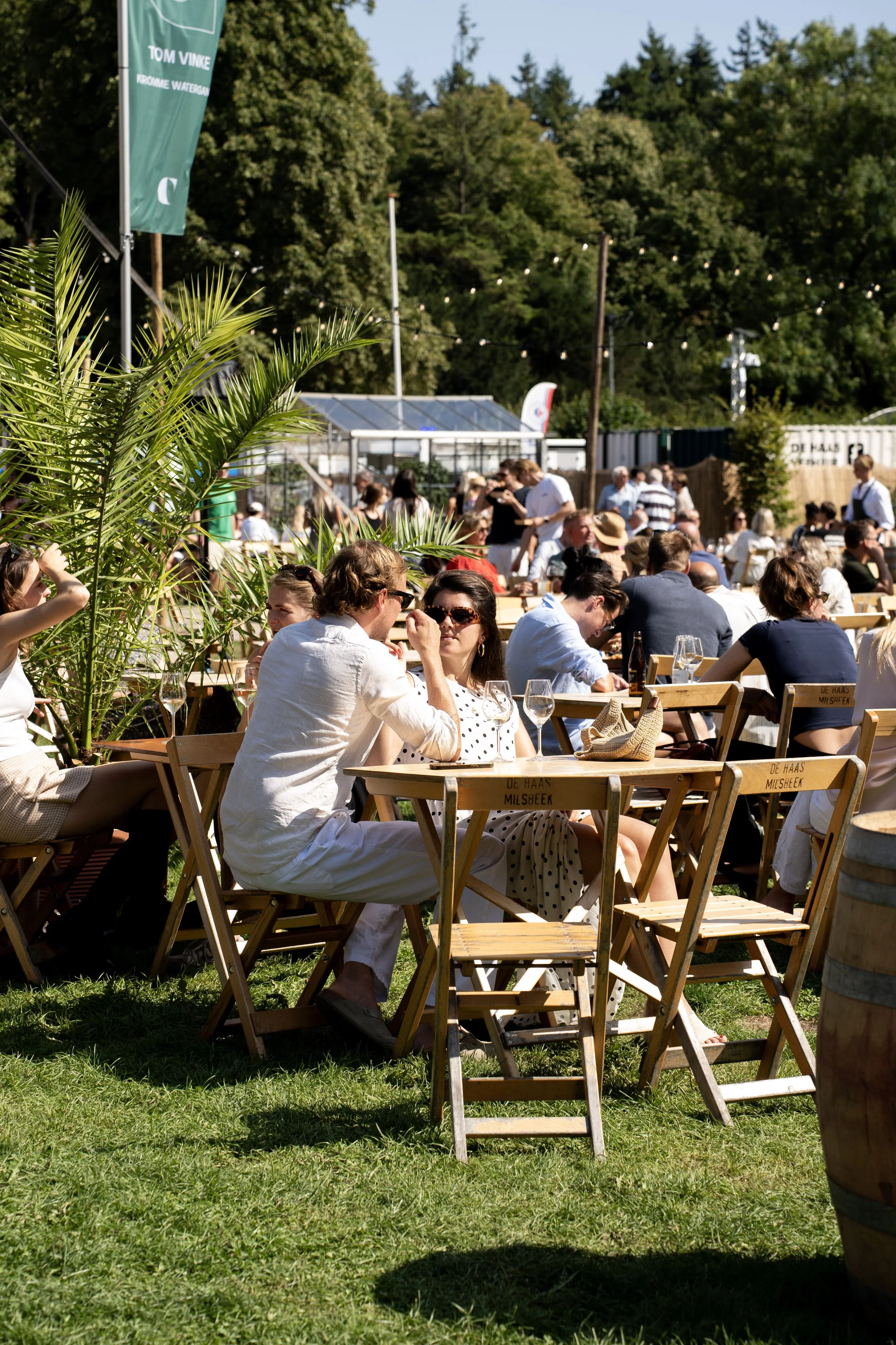 Een zomerse outdoor evenementenlocatie met mensen die aan tafels zitten, genietend van eten en drinken, met groene bomen en een blauwe lucht op de achtergrond.