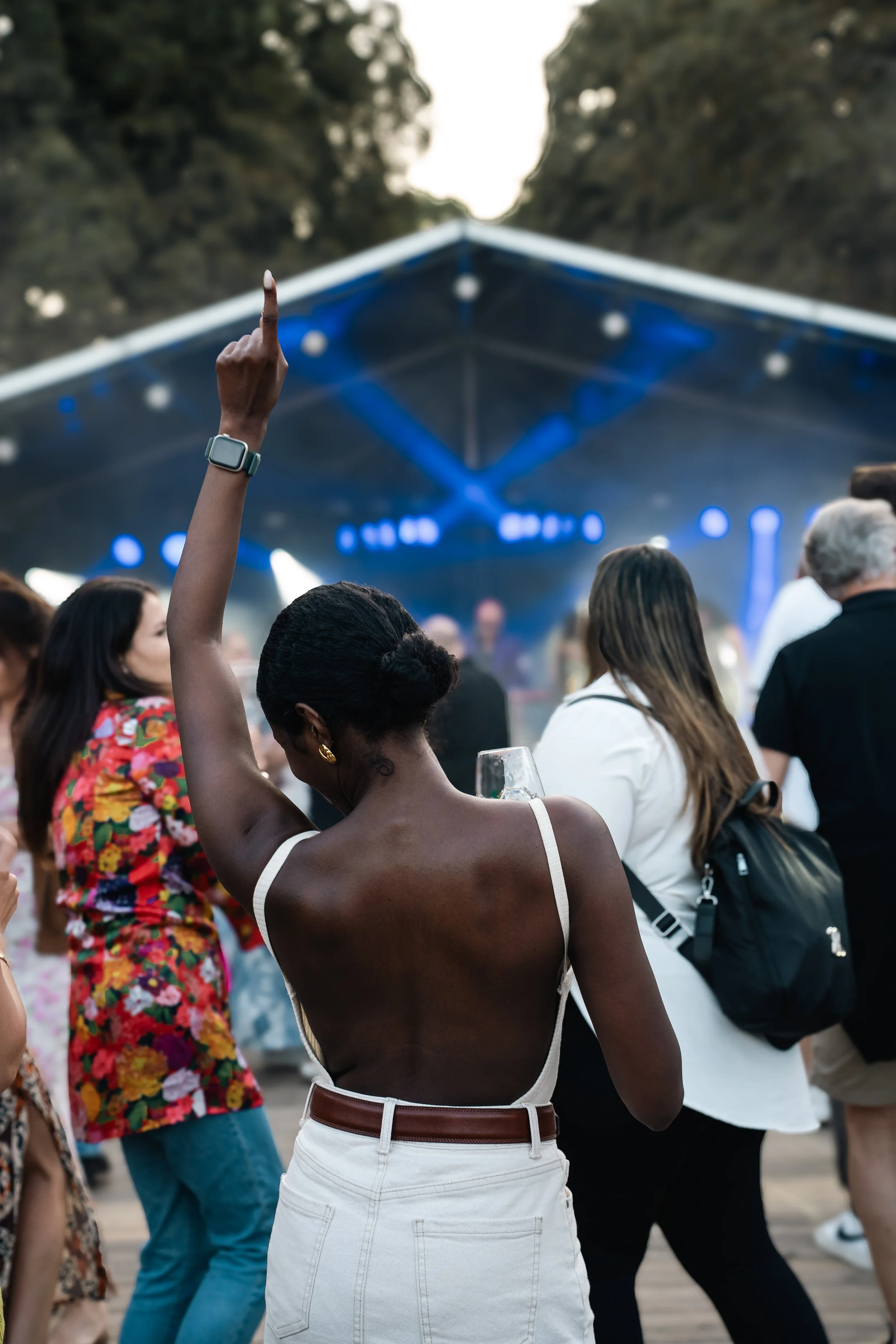 Publiek bij outdoor concert met een persoon die omhoog wijst, in de achtergrond een podium met blauwe verlichting.