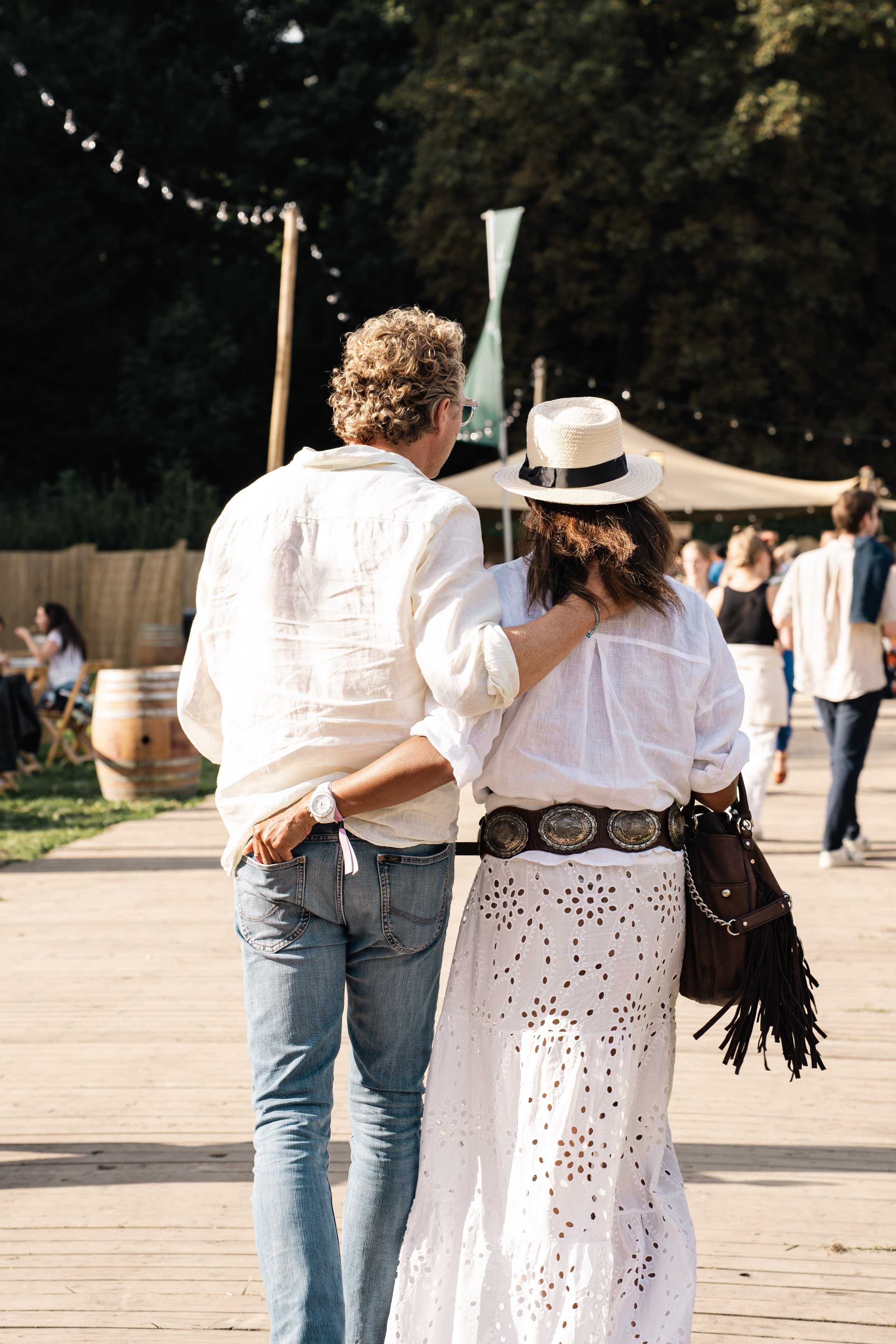 Twee mensen wandelen samen, een man en een vrouw. De man heeft krullend haar en draagt een witte blouse en jeans. De vrouw draagt een hoed met een zwarte band en een witte jurk met oogjespatroon. Ze lopen op een houten pad, met een menigte mensen op 