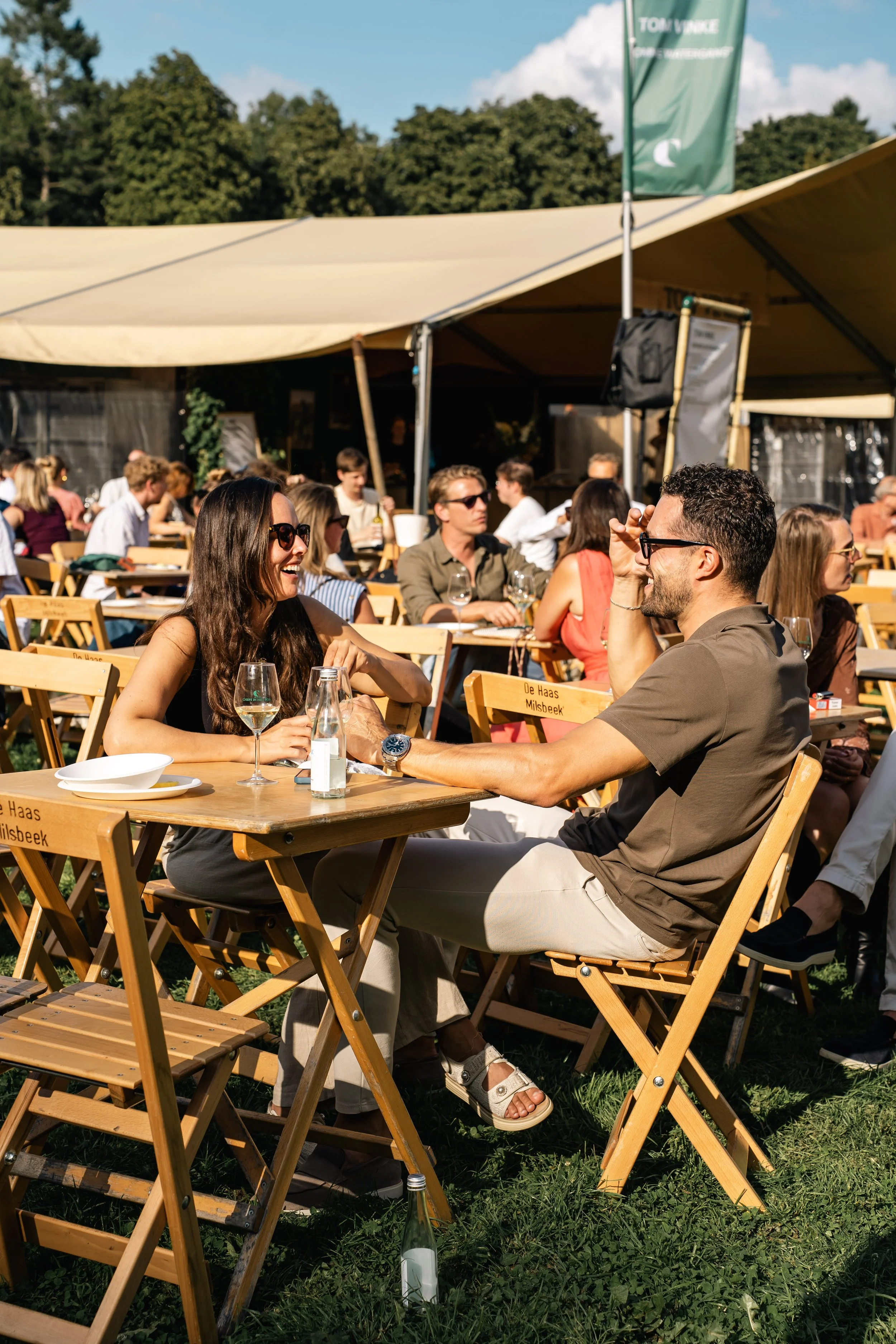 Mensen zitten in een zonnig buitenrestaurant onder een grote tent, lachend en pratend, met wijn en drinken op de tafels.