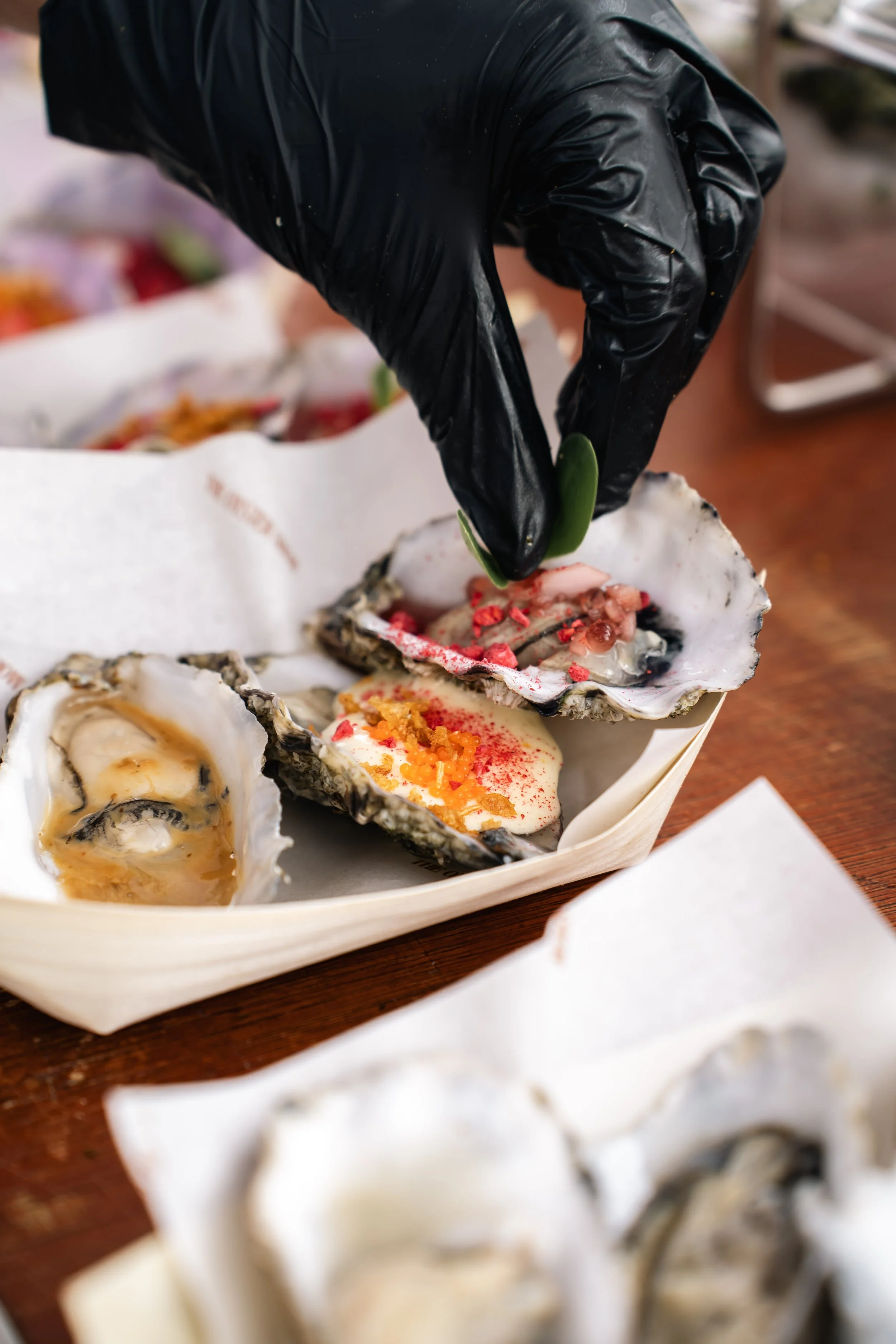 Close-up van een hand in een zwarte latex handschoen die een oester opent met een scalpel, met verschillende oesters in een houten bak.