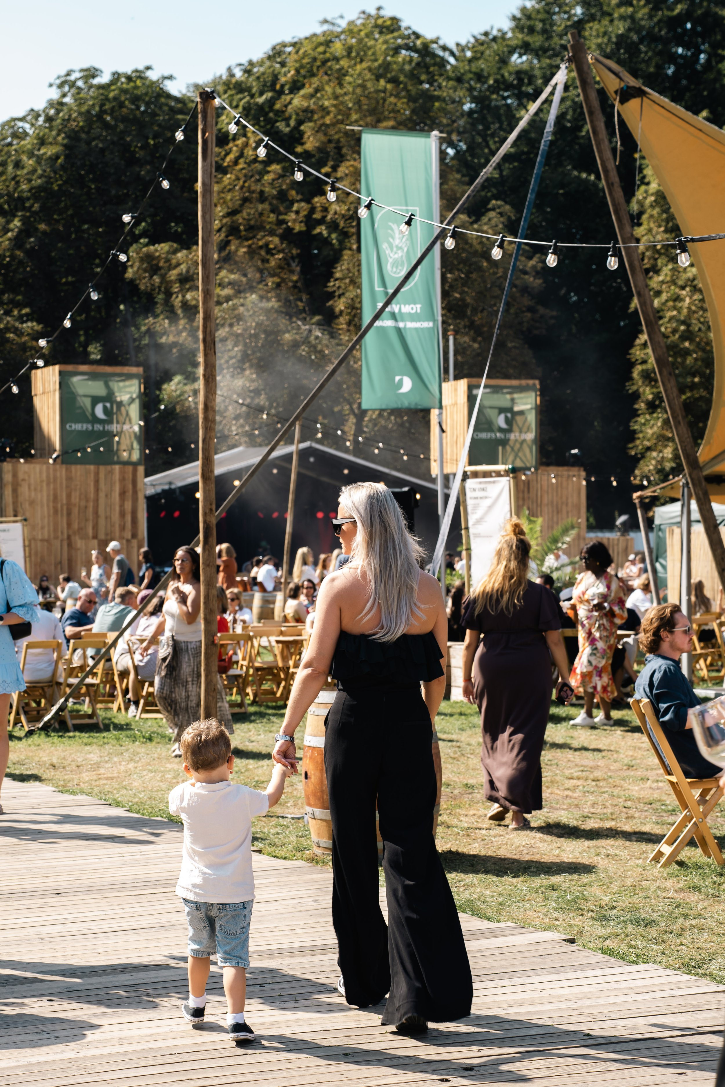 Mensen genieten buiten op een zonnige dag tijdens een festival of markt met zitplaatsen, houten paviljoens, en grote vlaggen, omgeven door bomen.