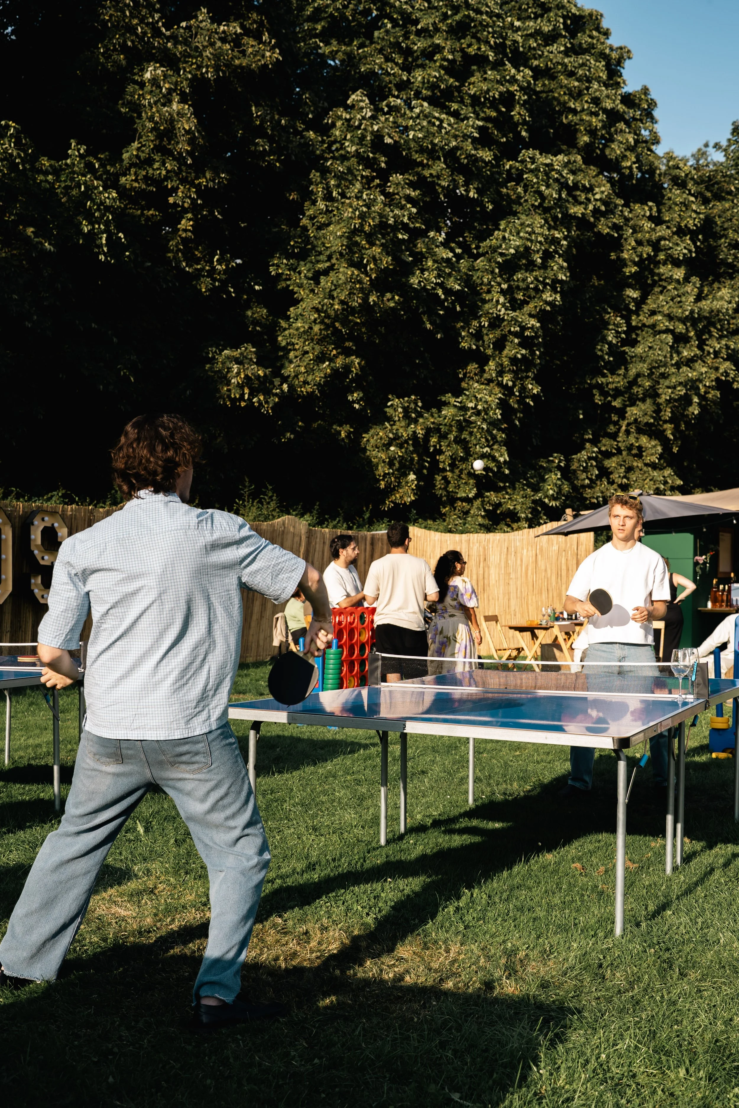 Mensen die buiten tafeltennis spelen en socializen in een tuin met een grote boom en een houten omheining.