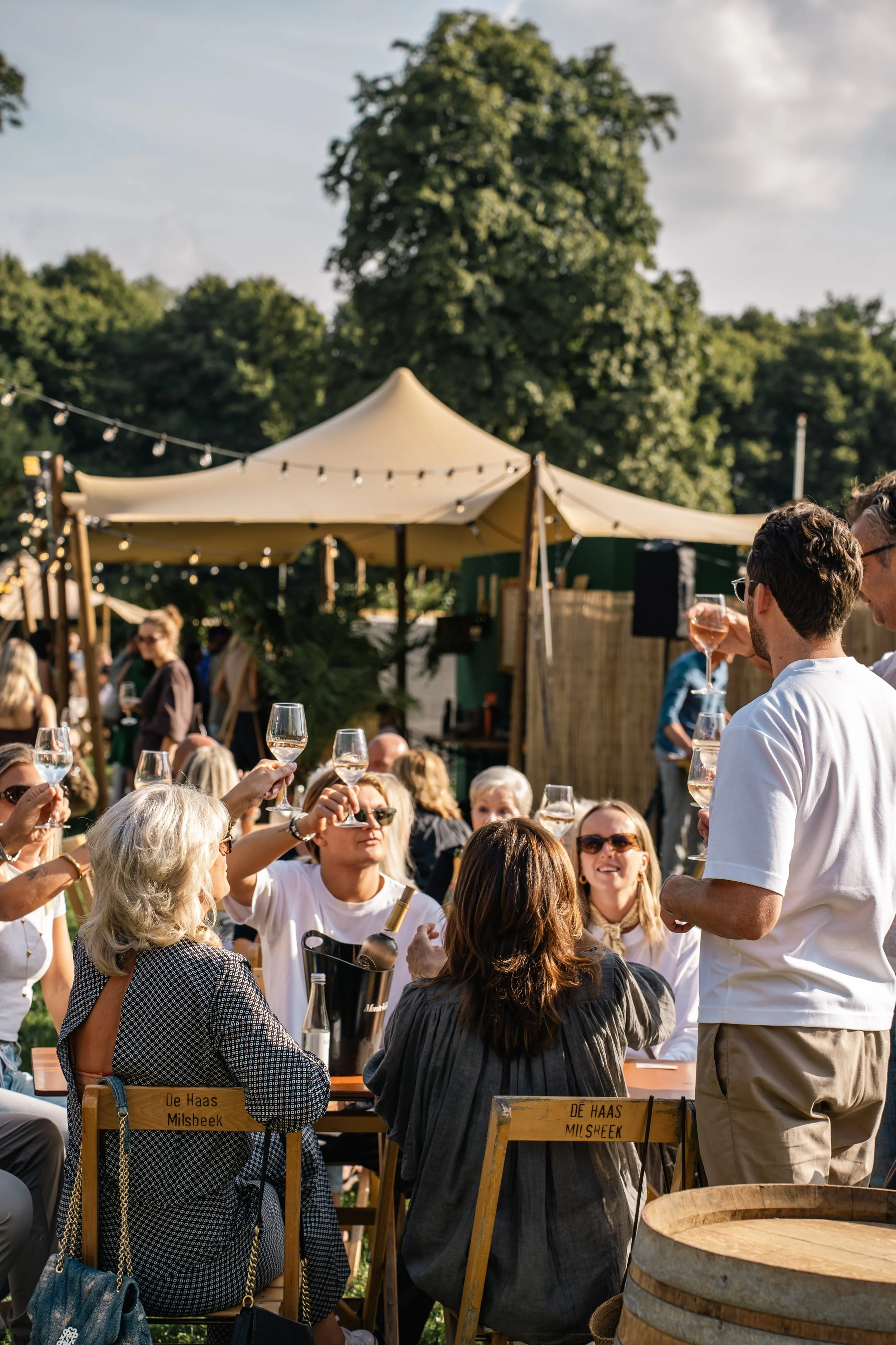 Mensen genieten van een drankje tijdens een buitenfeest onder een grote tent met lichtslingers. Er zitten mensen op houten banken met borden en glazen, op de achtergrond groene bomen en een blauwe hemel.