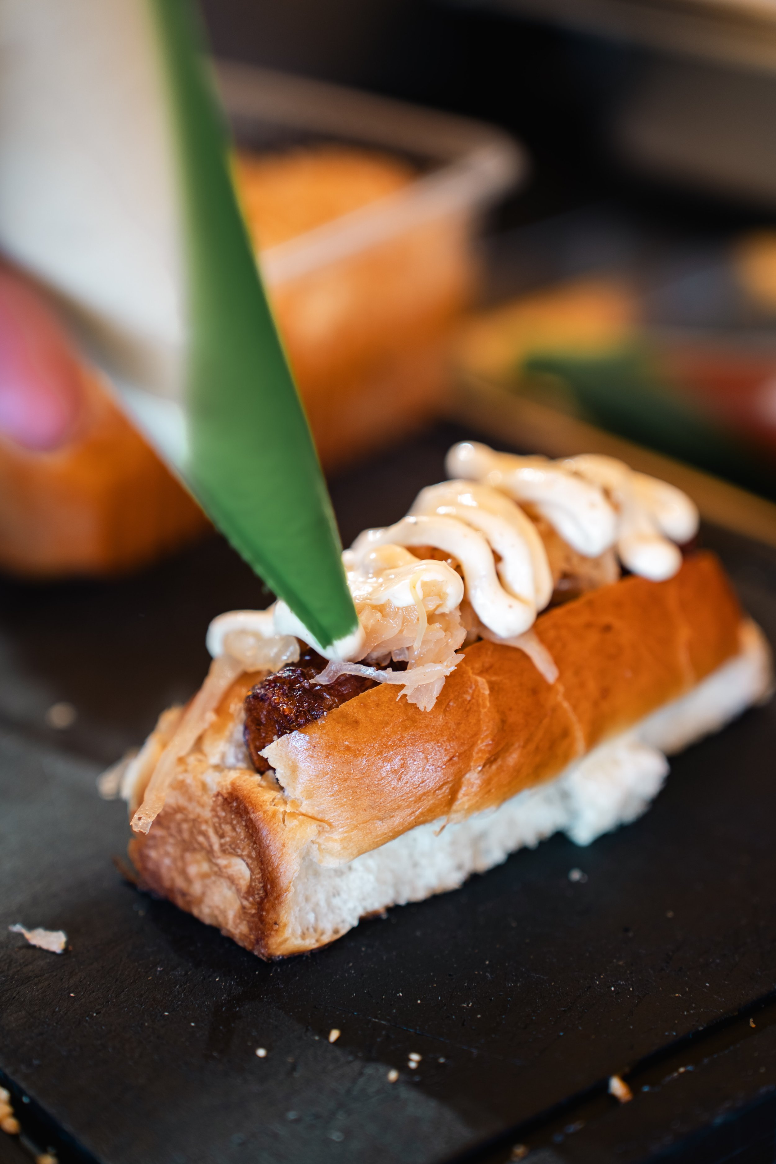 Een knapperige broodje met pulled pork, saus, en wortels, versierd met mayonaise en een groen blad, geserveerd op een zwarte plank.