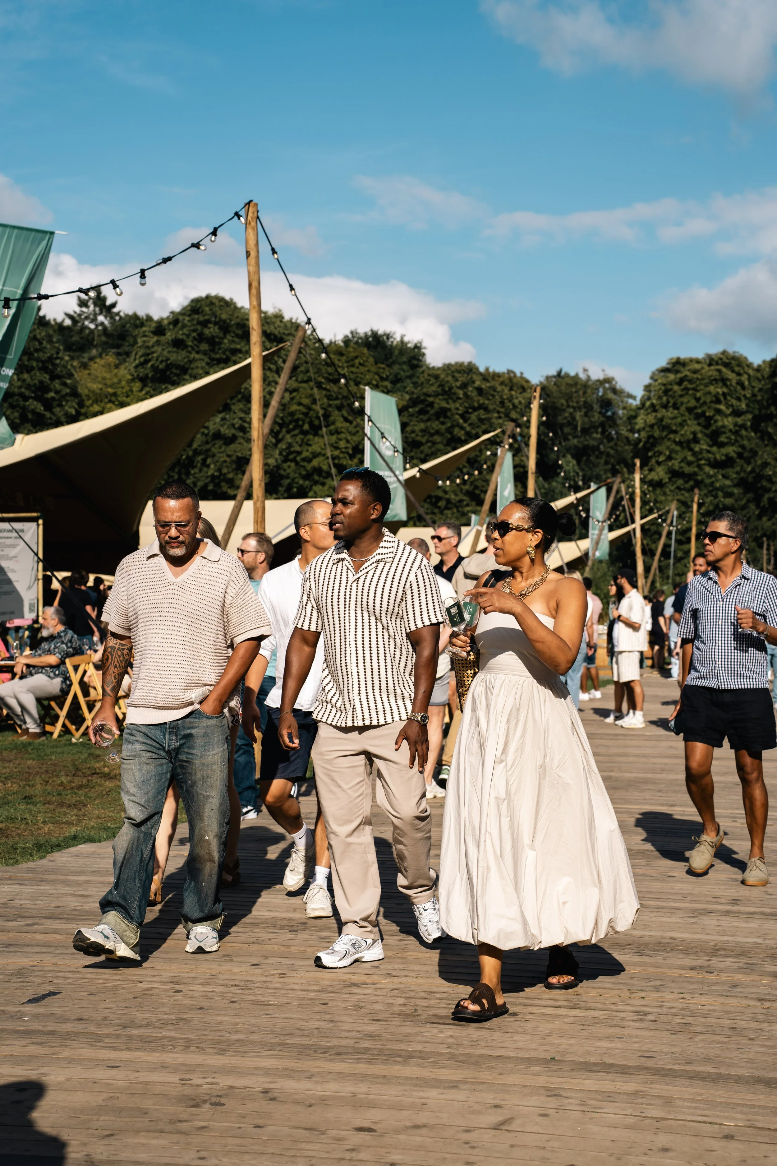 Mensen wandelen op een houten pad in een openluchtfestival, met tenten en bomen op de achtergrond en een blauwe lucht met wolken boven.