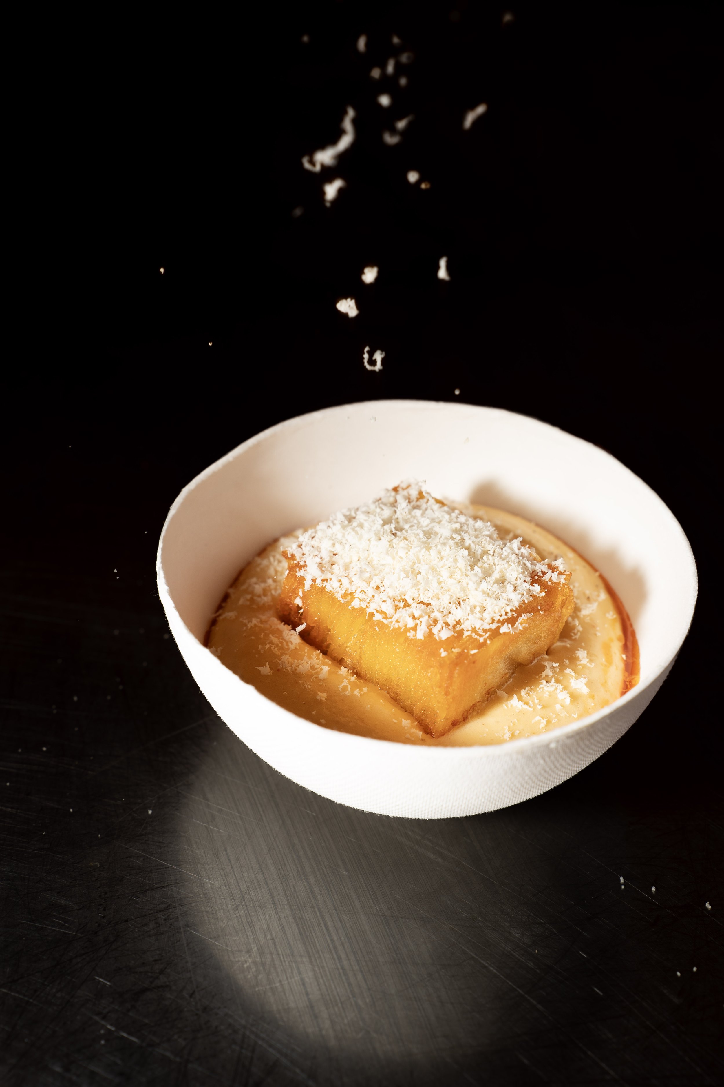 Een dessert in een witte kom met een krokant kussentje bovenop, bestrooid met geraspte kokos, op een zwarte tafel.