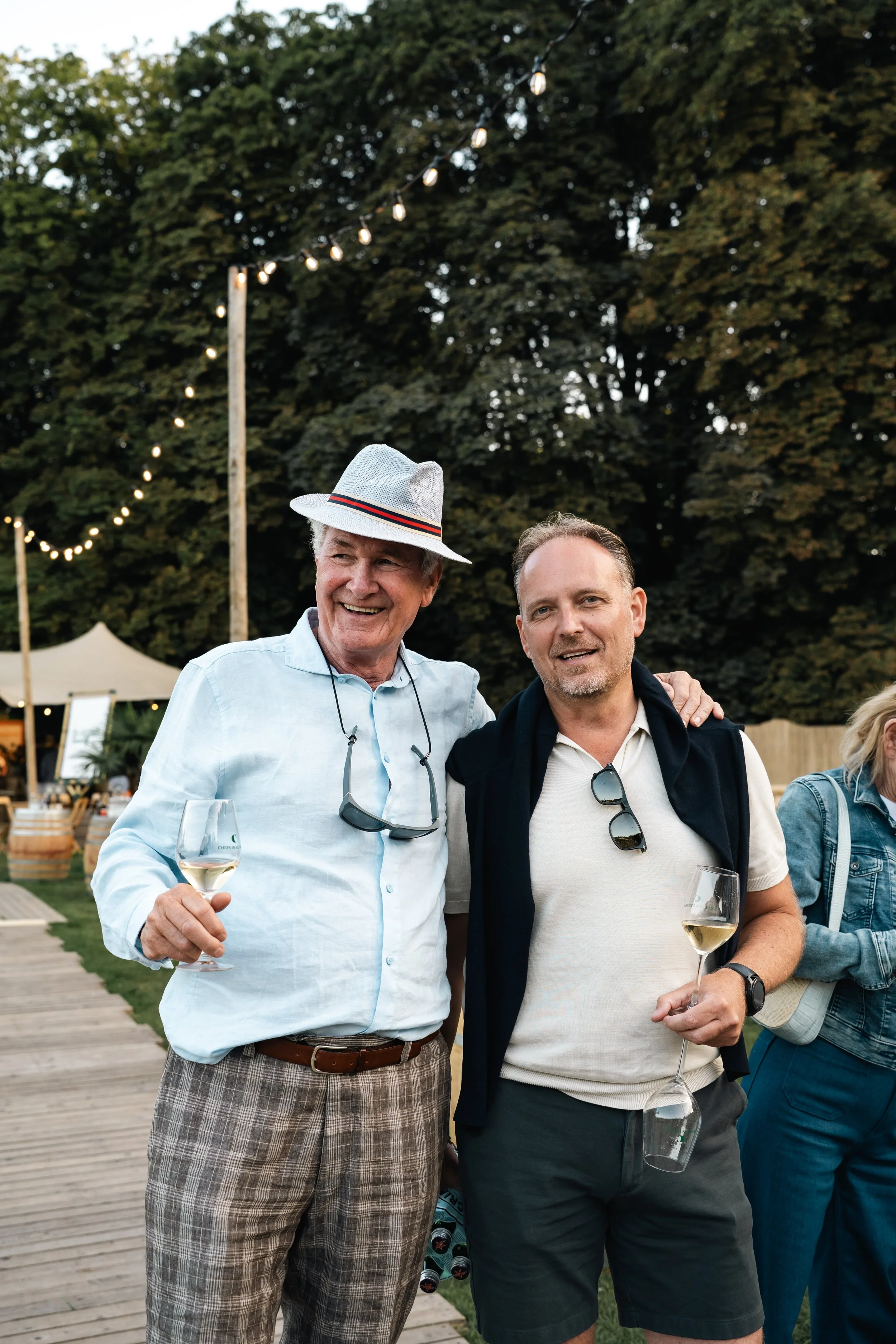 Twee mannen met glazen witte wijn, glimlachend en knuffelend op een buitenevenement met verlichting en groene bomen op de achtergrond.