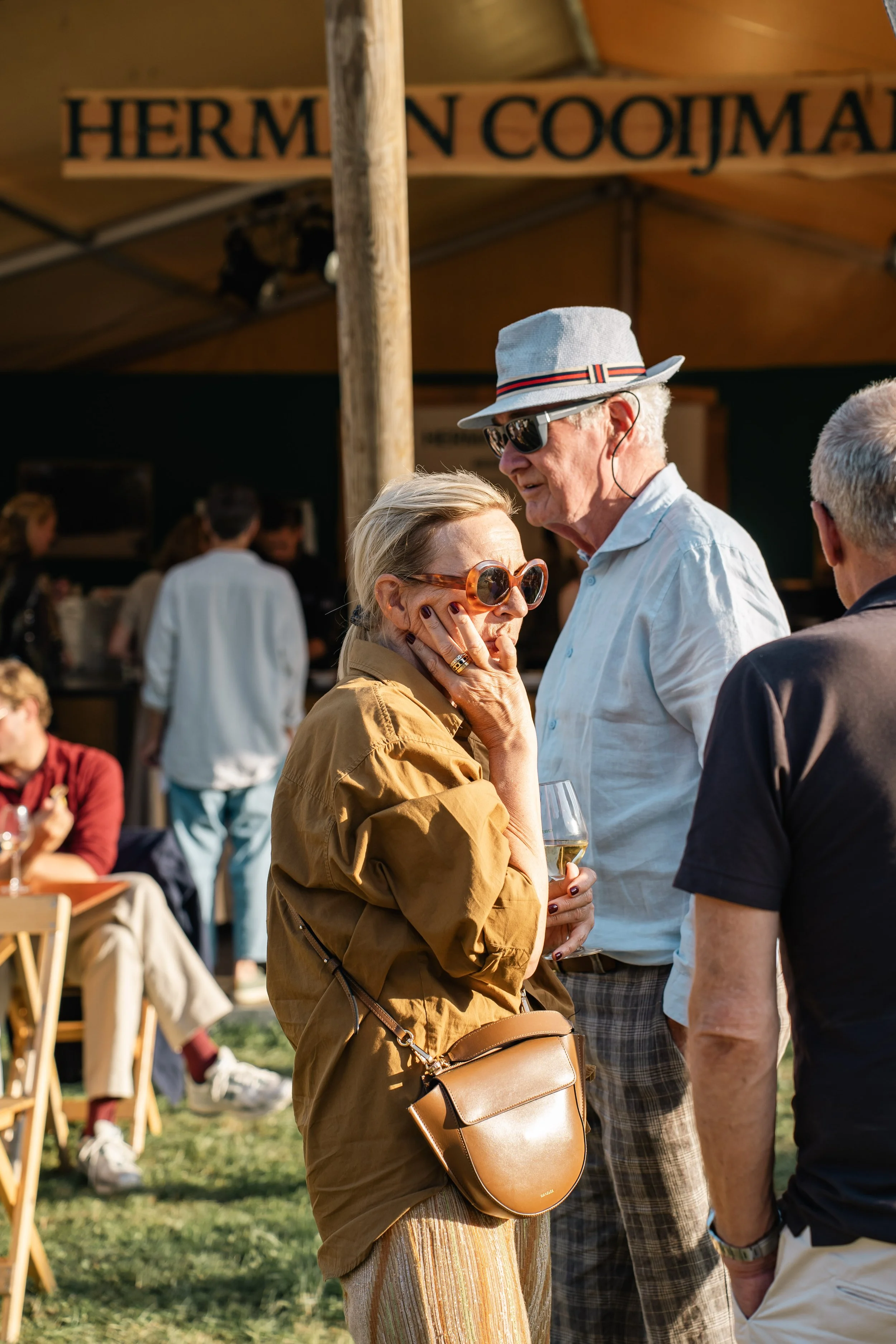 Jongens en meisjes op een terras in de zon. Ze genieten van een drankje en gesprekken. Vrouwen en mannen spelen en praten in de achtergrond. Er hangt een informele sfeer.