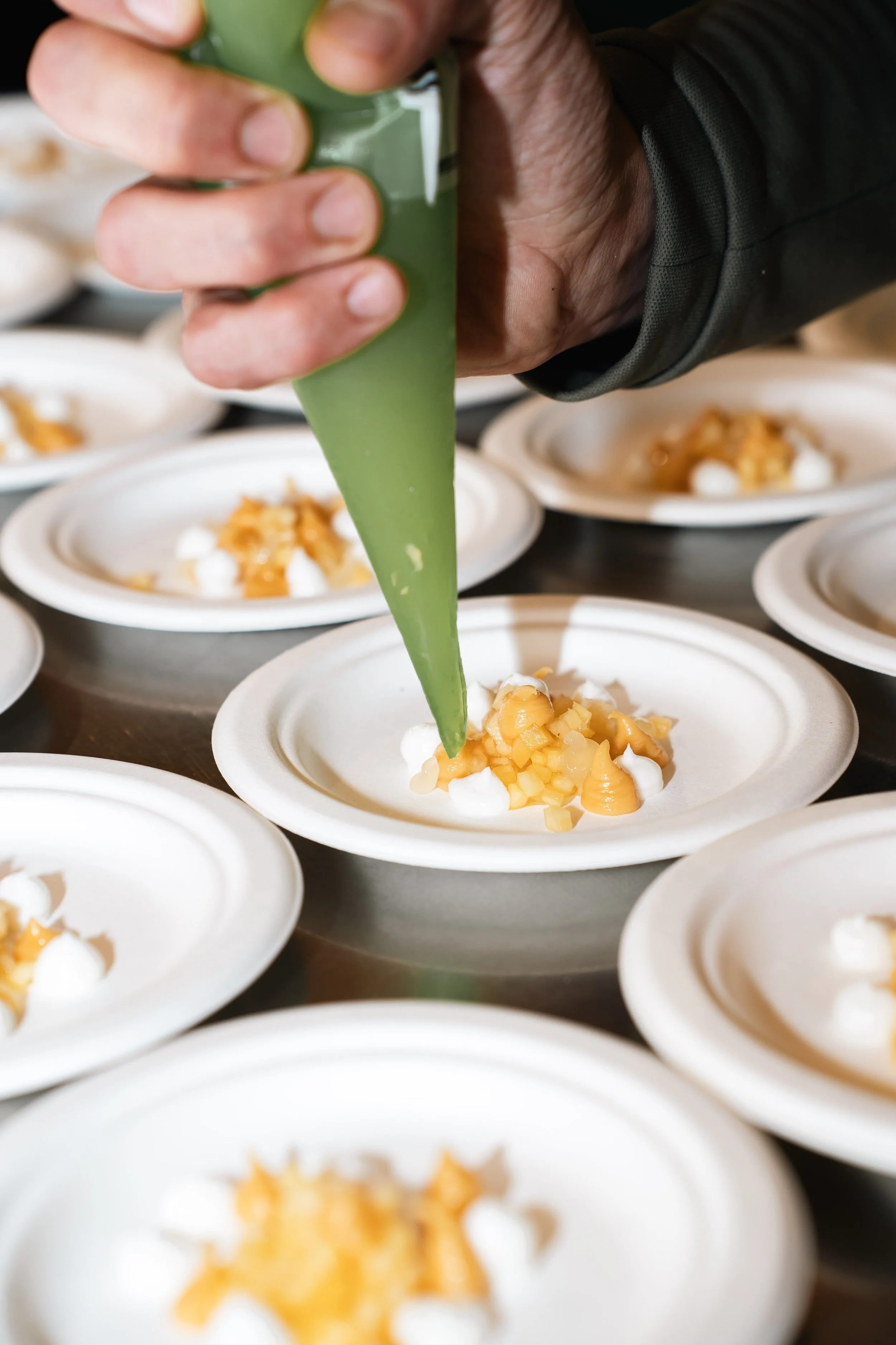 Persoon die groene saus uit een tube op een dessert schept, met meerdere schaaltjes met dessert met ijs en fruit op de achtergrond.