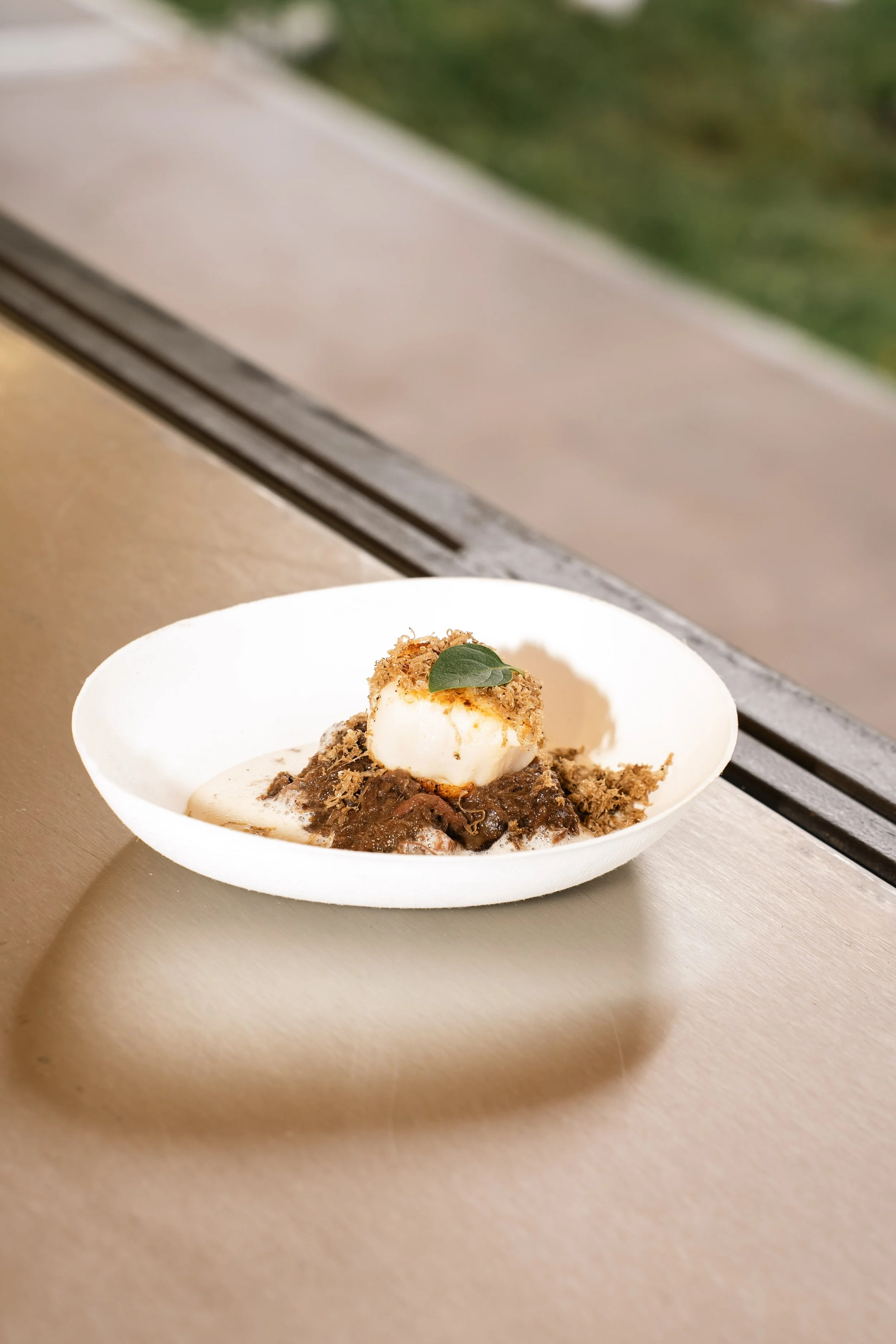 Een wit bord met een gerecht bestaande uit vlees, een schepje mayonaise, een gepaneerde kipfilet met een blaadje basilicum, geserveerd op een houten tafel met een raam op de achtergrond.