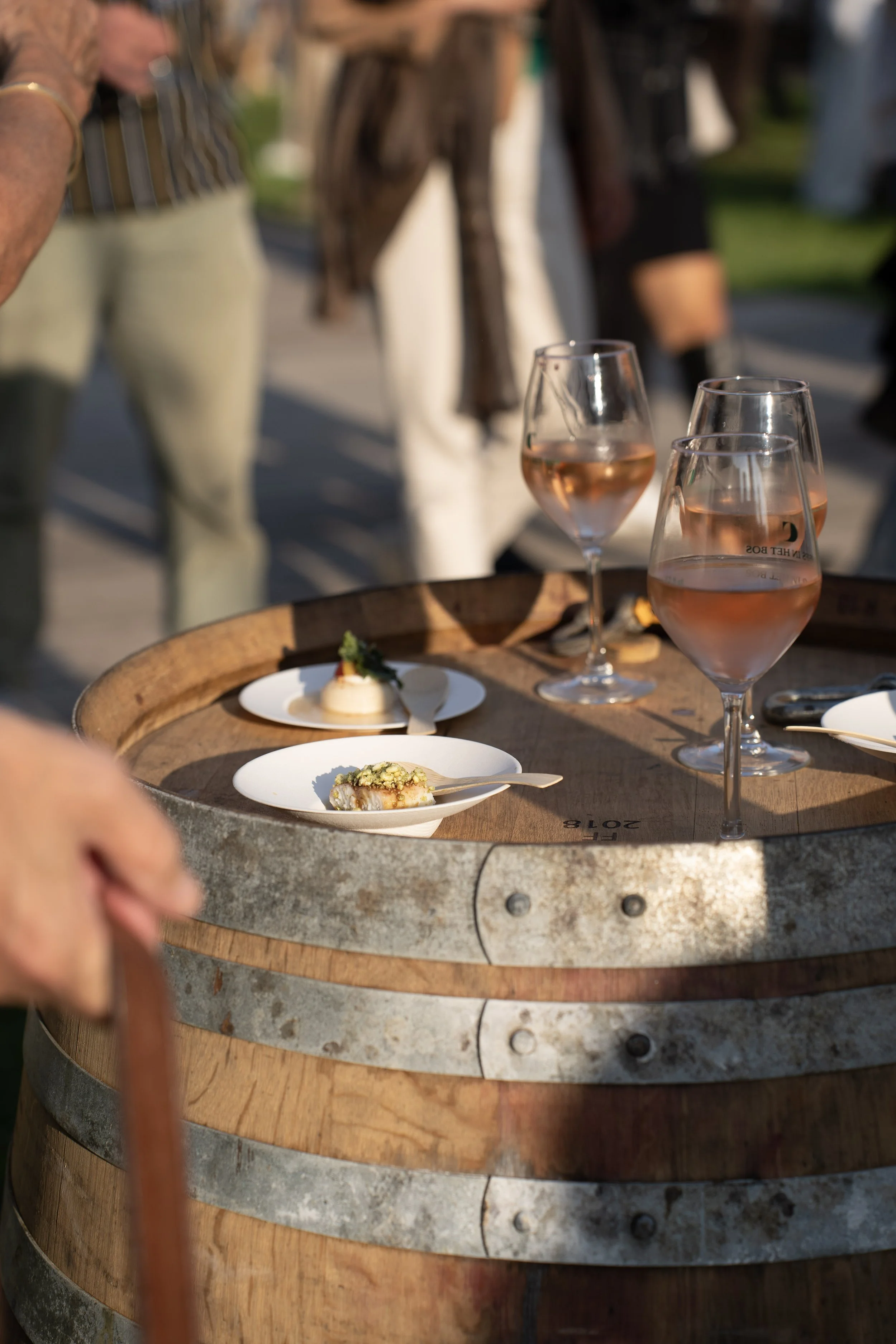 Drie glazen rosé wijn en twee kleine borden met hapjes op een houten wijnvat, in een buitenomgeving met mensen op de achtergrond.