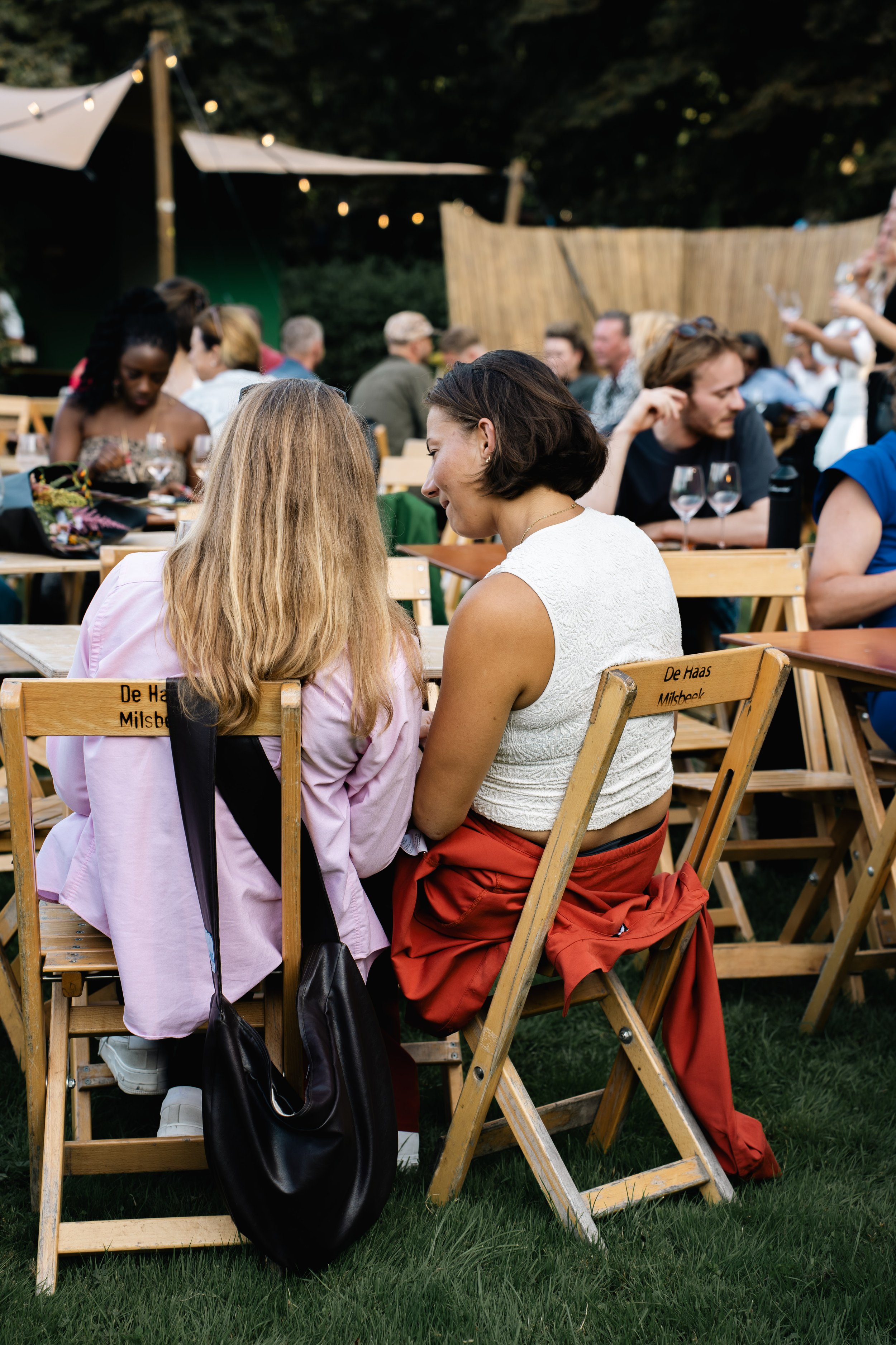 Twee vrouwen die met hun hoofden dicht tegen elkaar zitten, op een houten stoel tijdens een buitenfeest, rond een houten tafel.