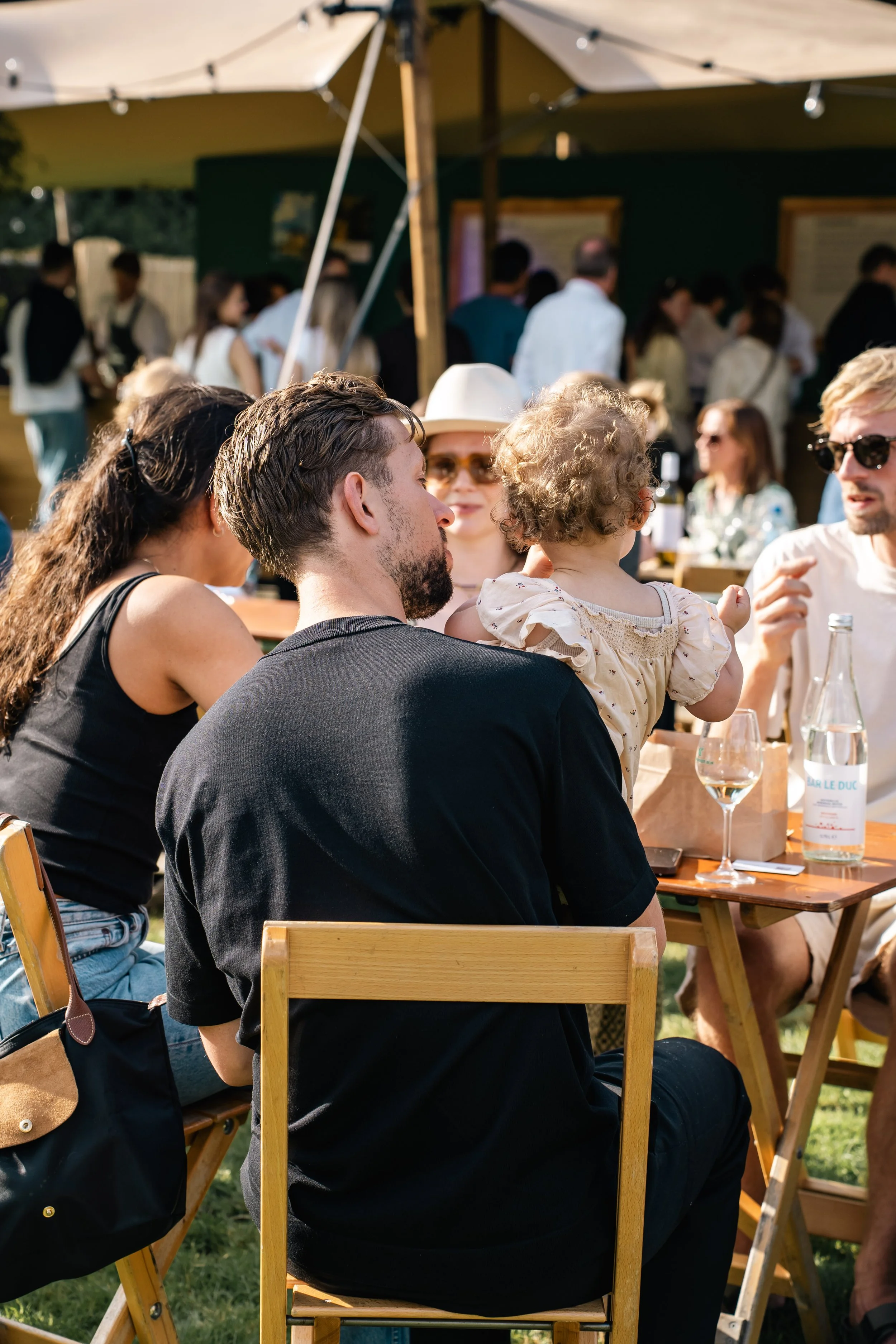 Mensen zitten in een groep buiten tijdens een evenement, onder een parasol, met drankjes op tafel, en een menigte op de achtergrond.
