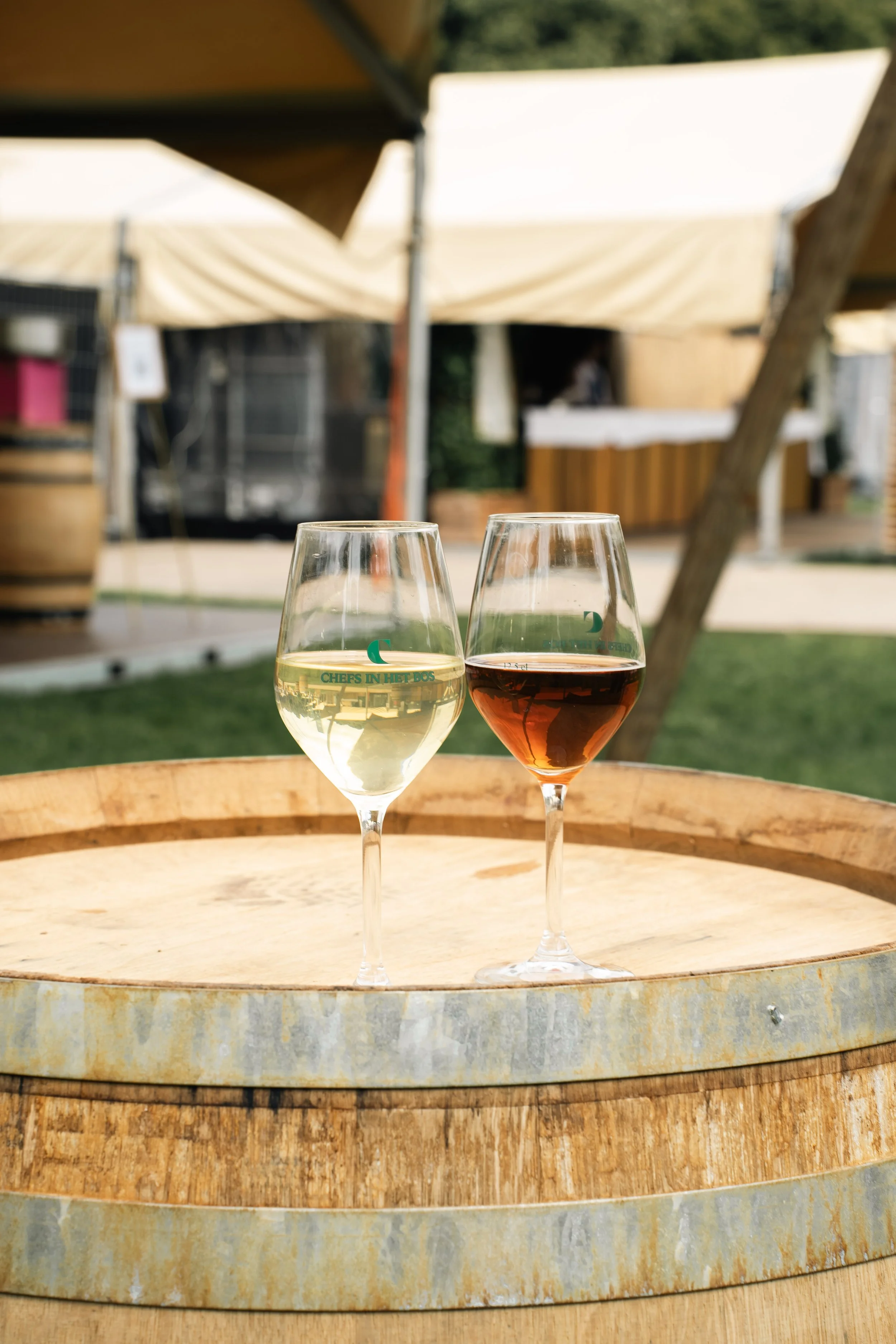 Twee wijnglazen, één met witte wijn en één met rosé, op een houten vat onder een buitenparasol, met een pot en een tent in de achtergrond.