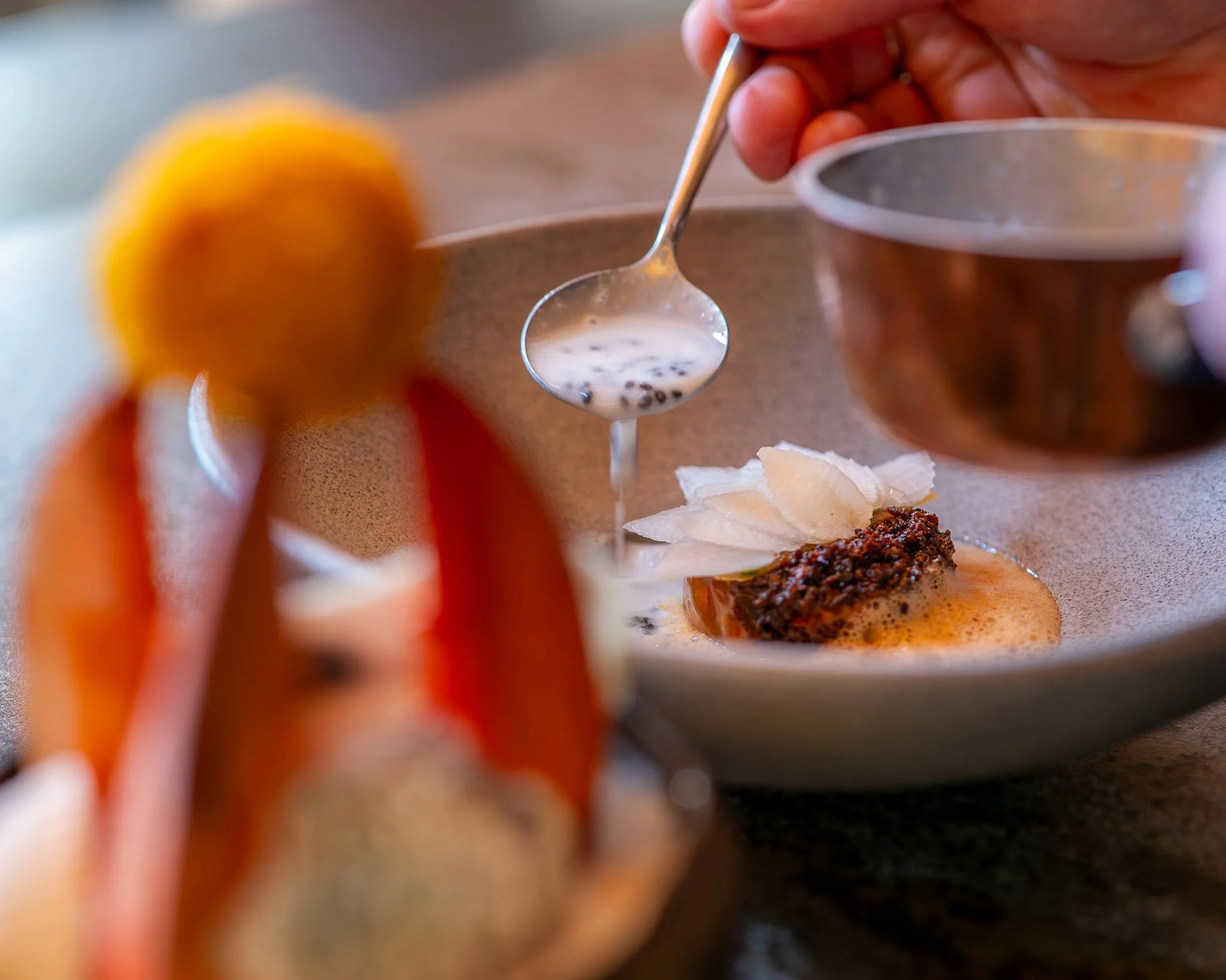 Een zintuiglijke close-up van een innovatief gerecht waarbij een rijke saus wordt geschonken, perfect passend bij het culinaire Roots of 2050 thema op het outdoor foodfestival Chefs in het Bos.