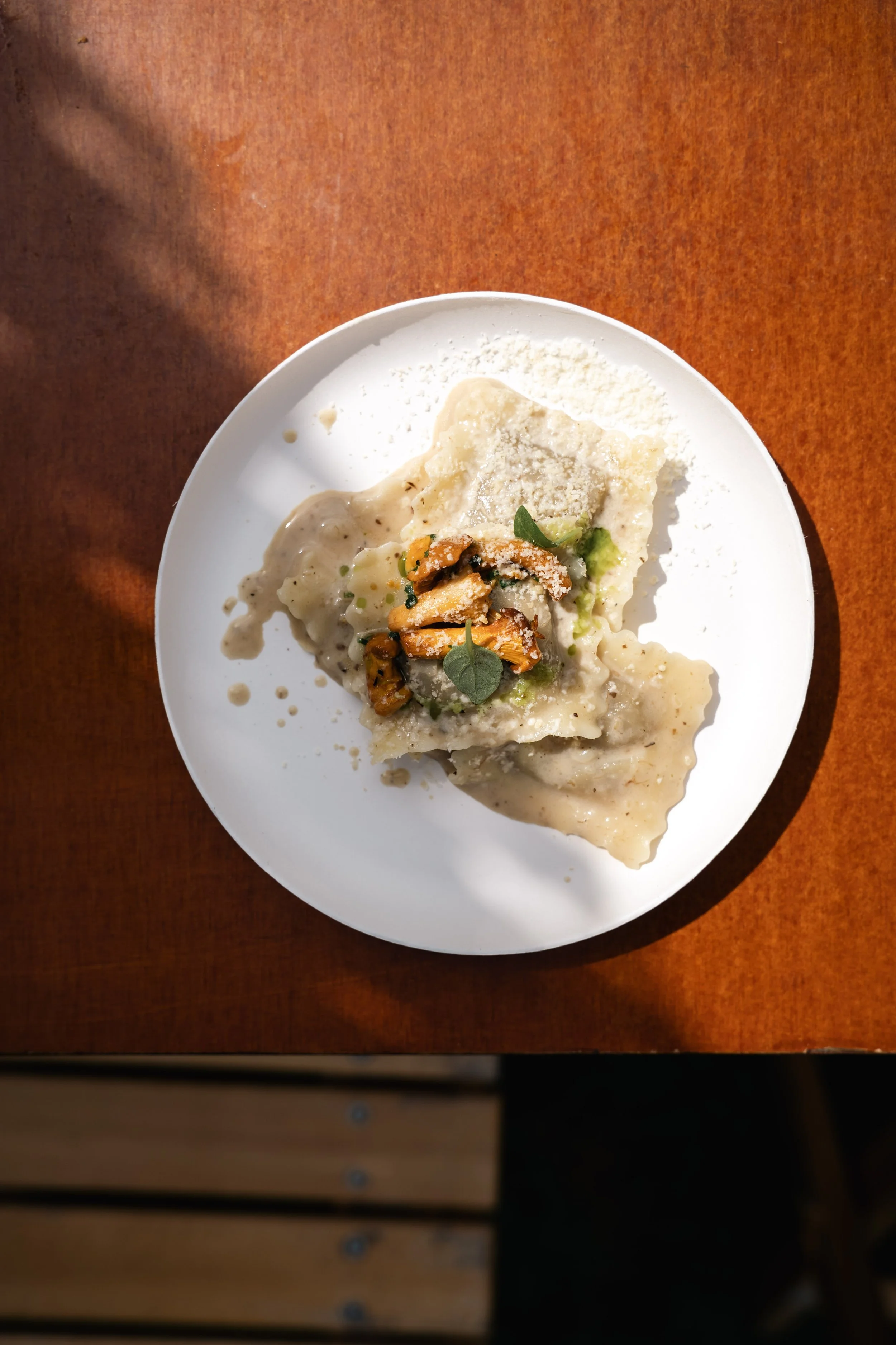 Het gerecht is ravioli met een romige saus, garnering met een liefdevol geplaatste groene bladkruid en gekaramelliseerde pompoenstukjes, geserveerd op een witte bord op een houten tafel.