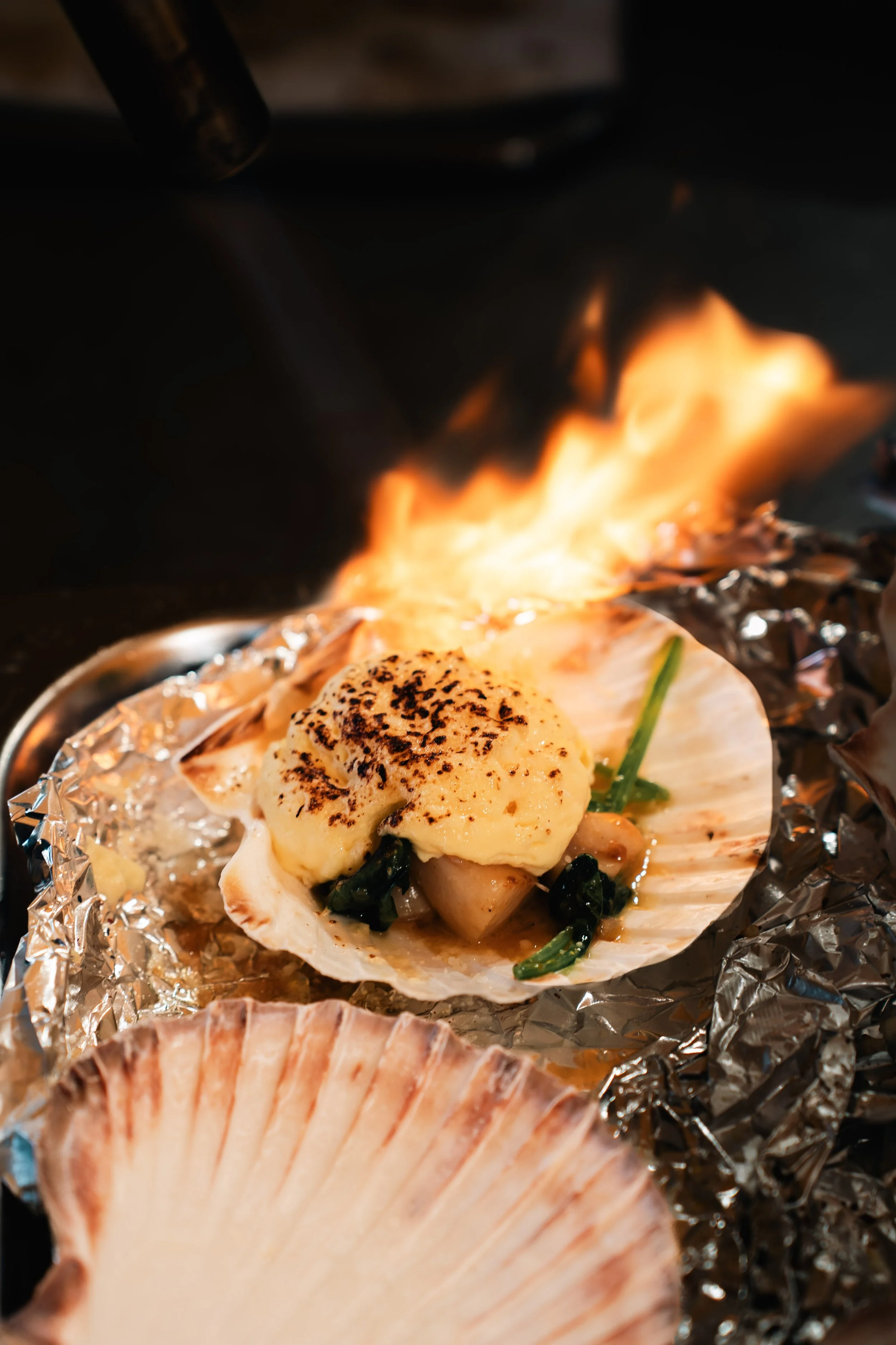 Geroosterde coquilles met kruiden en groenten op aluminiumfolie, brandende vlammen in achtergrond.