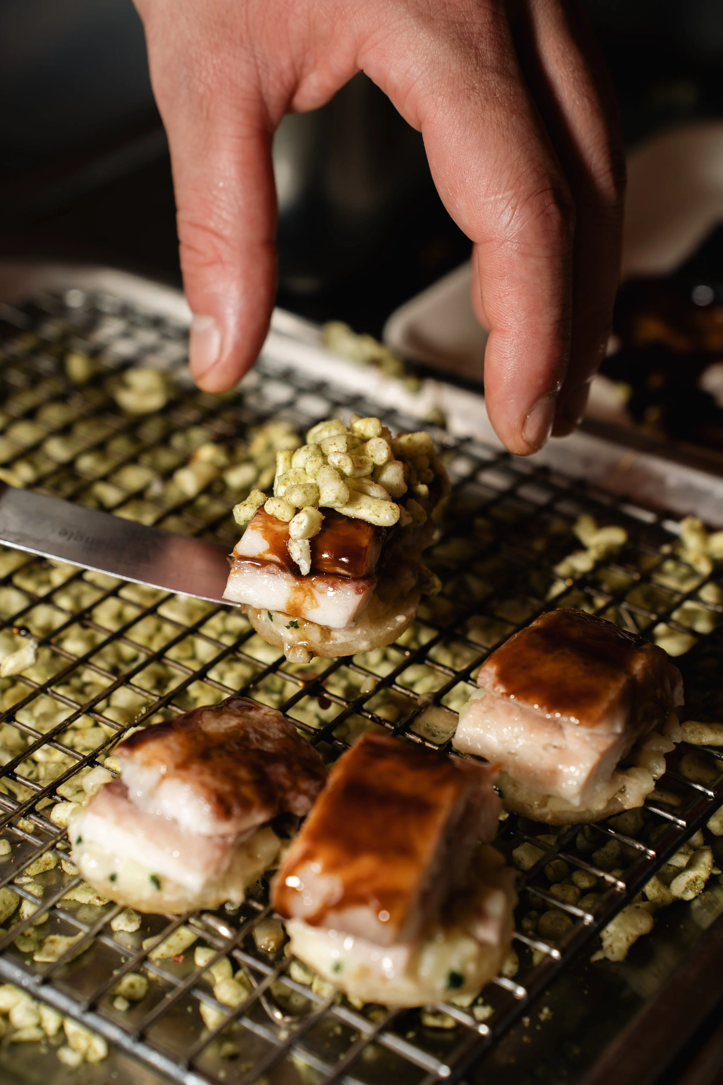 Close-up van een hand die sushi met topping vastpakt boven een rooster met stukjes vis en saus.