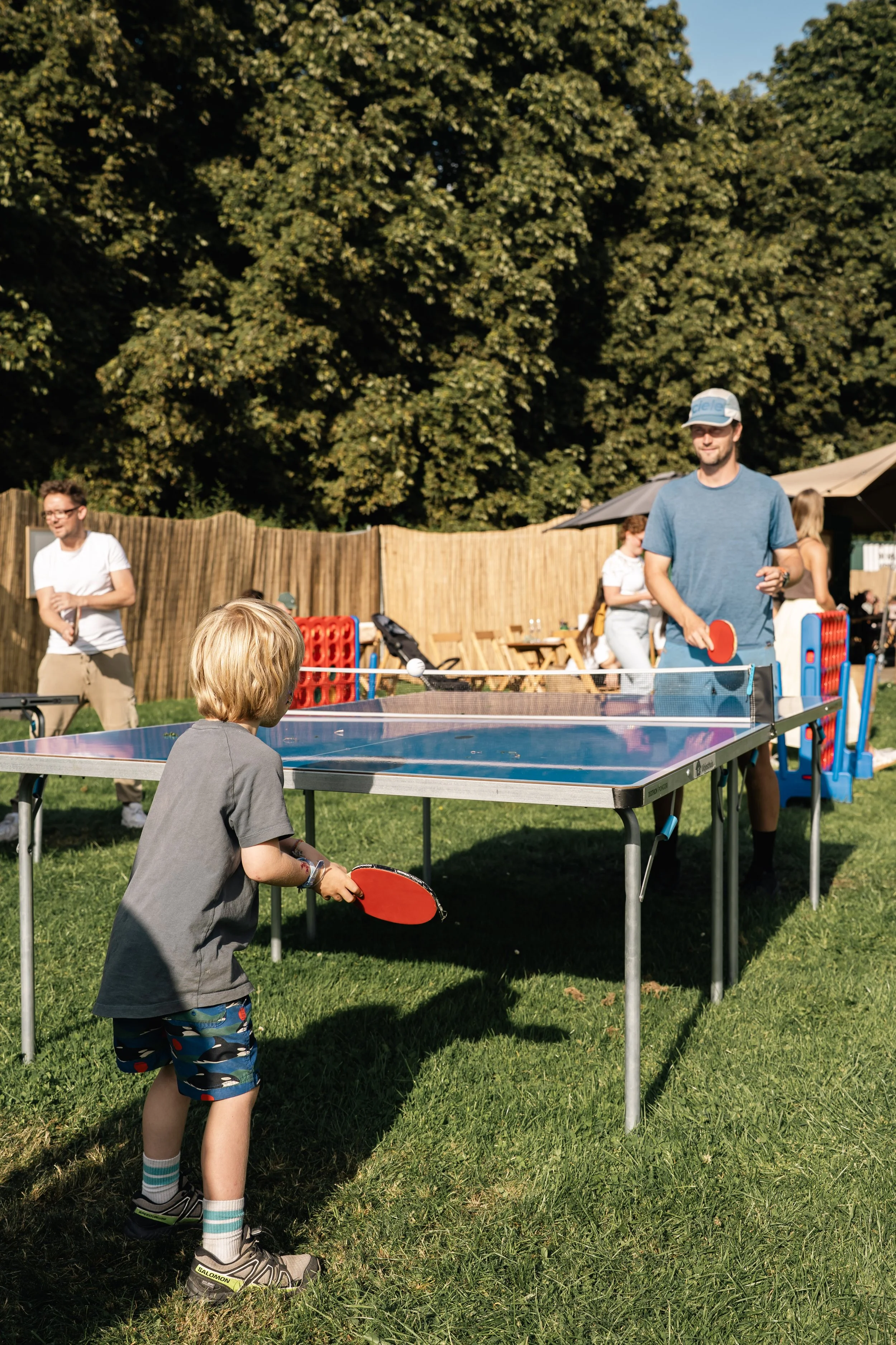 Kind speelt tafeltennis met volwassen in een tuin met groen gras en grote bomen, omgeven door een houten hek en parasols.