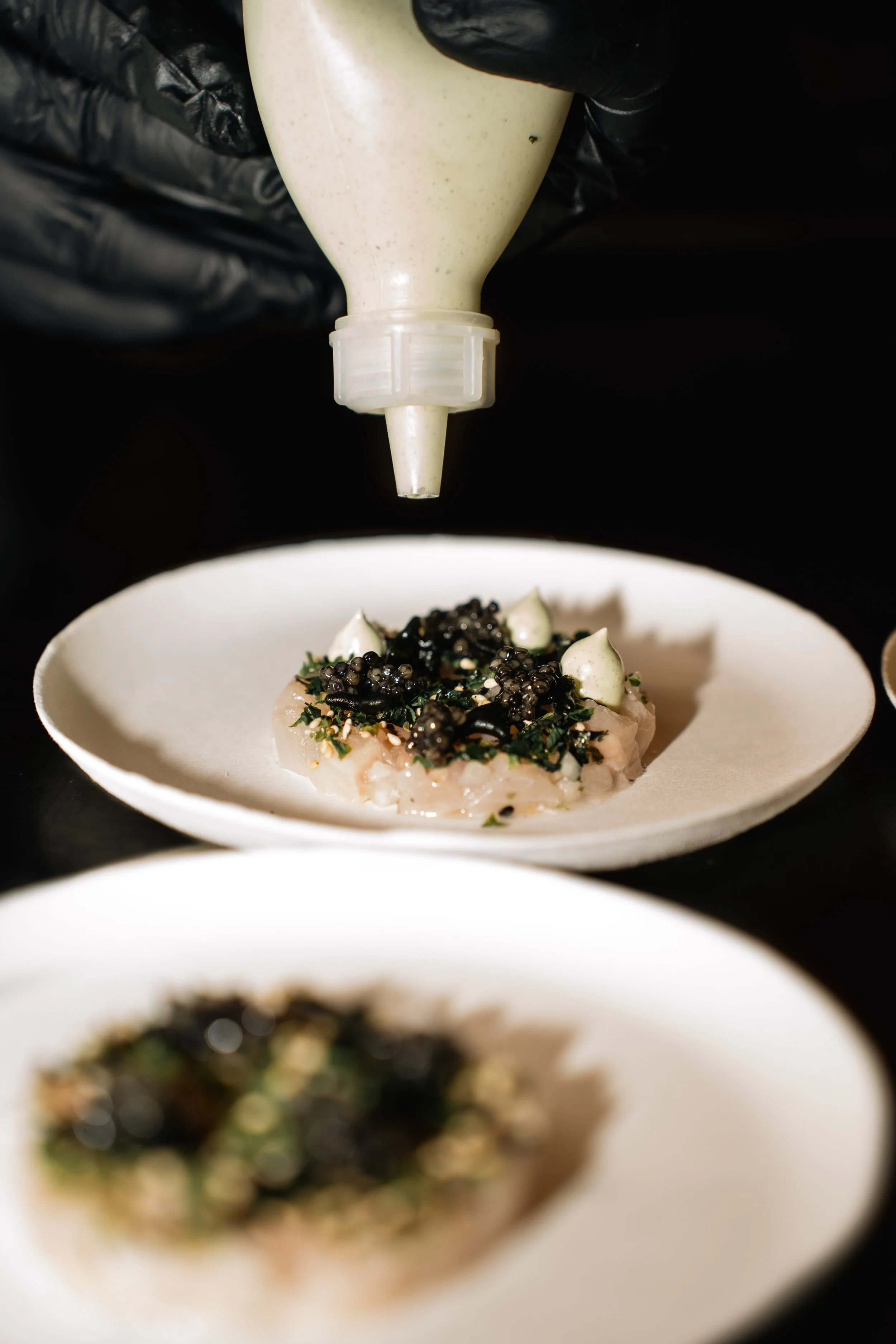 Een chef-kok spuit saus of topping over een gerecht met groenten en caviarlike pepers in een witte kom, de achtergrond is zwart.