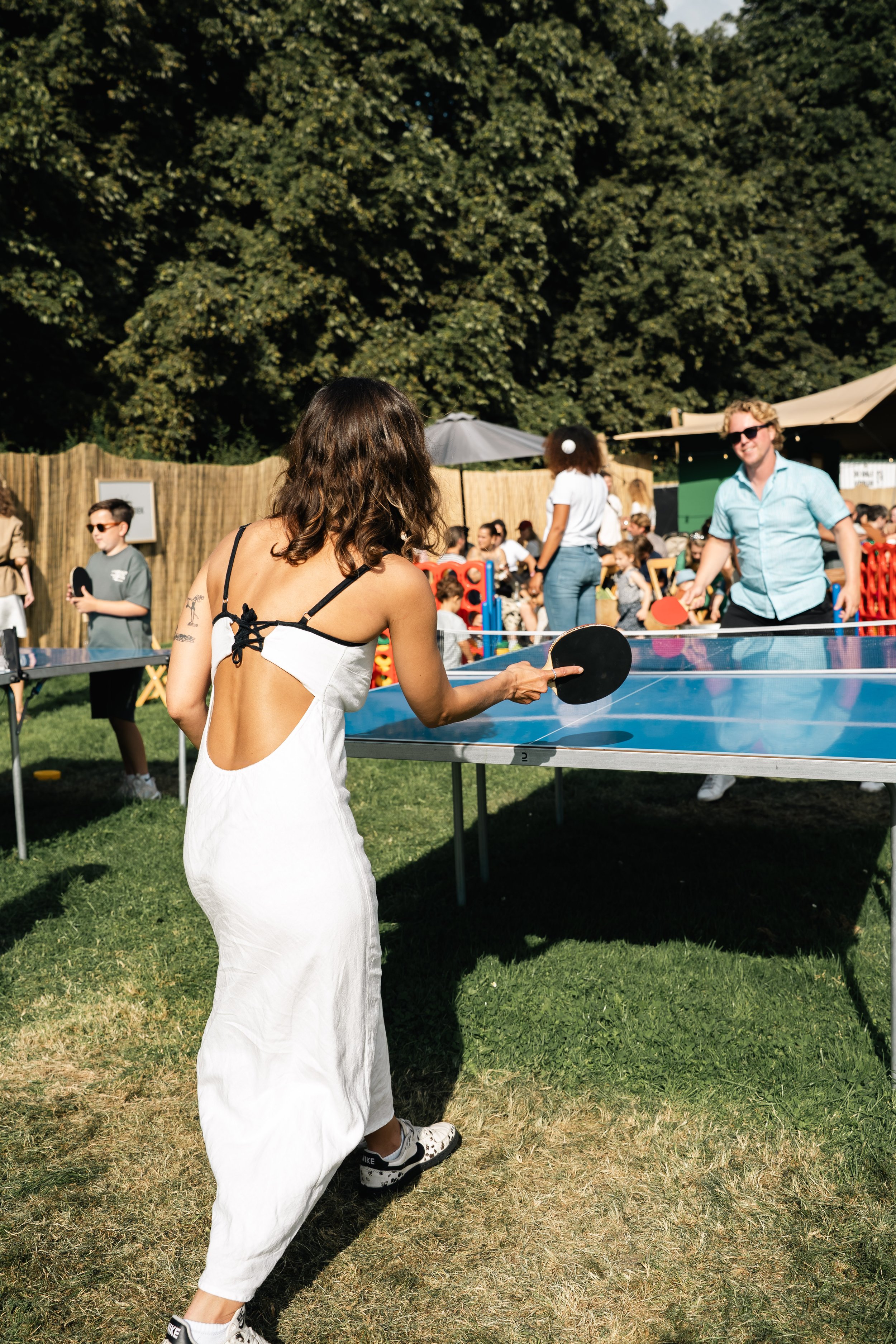 Een vrouw in een witte jurk die pingpong speelt op een grasveld tijdens een zomers evenement met veel mensen, bakstenen muur, parasols en groene bomen op de achtergrond.