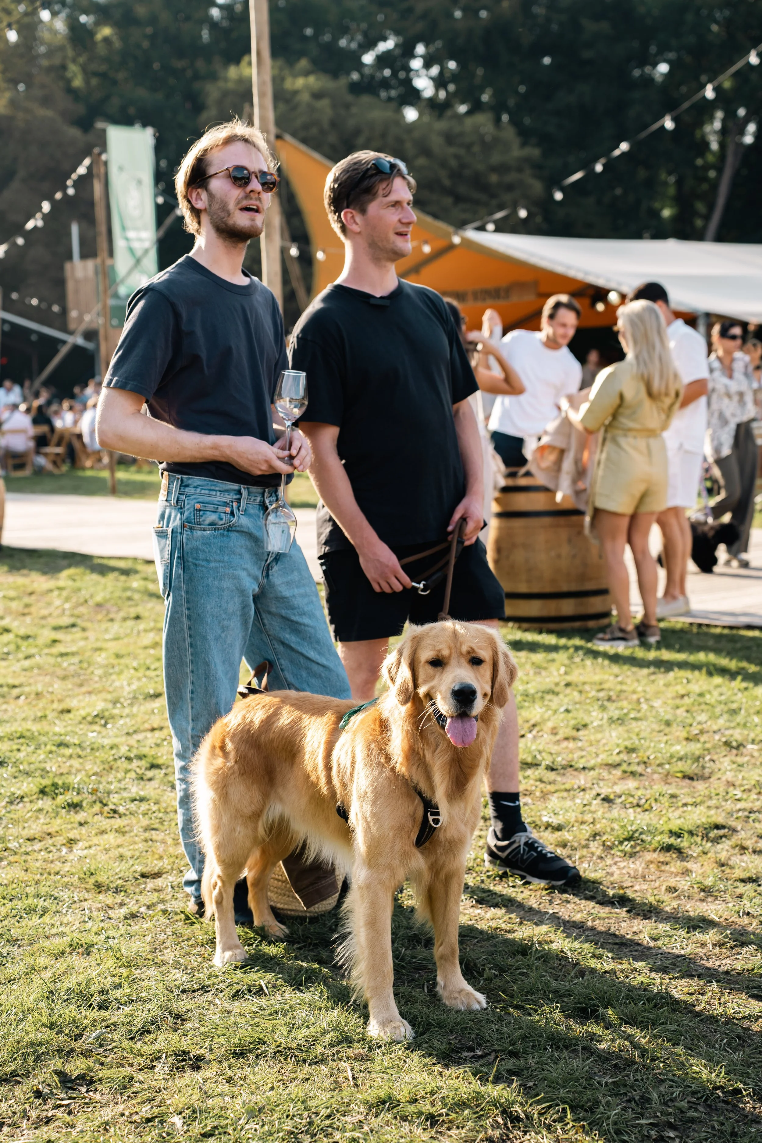 Twee jonge mannen met een hond en een fles wijn in een park tijdens een festival, met mensen op de achtergrond onder een feesttent.