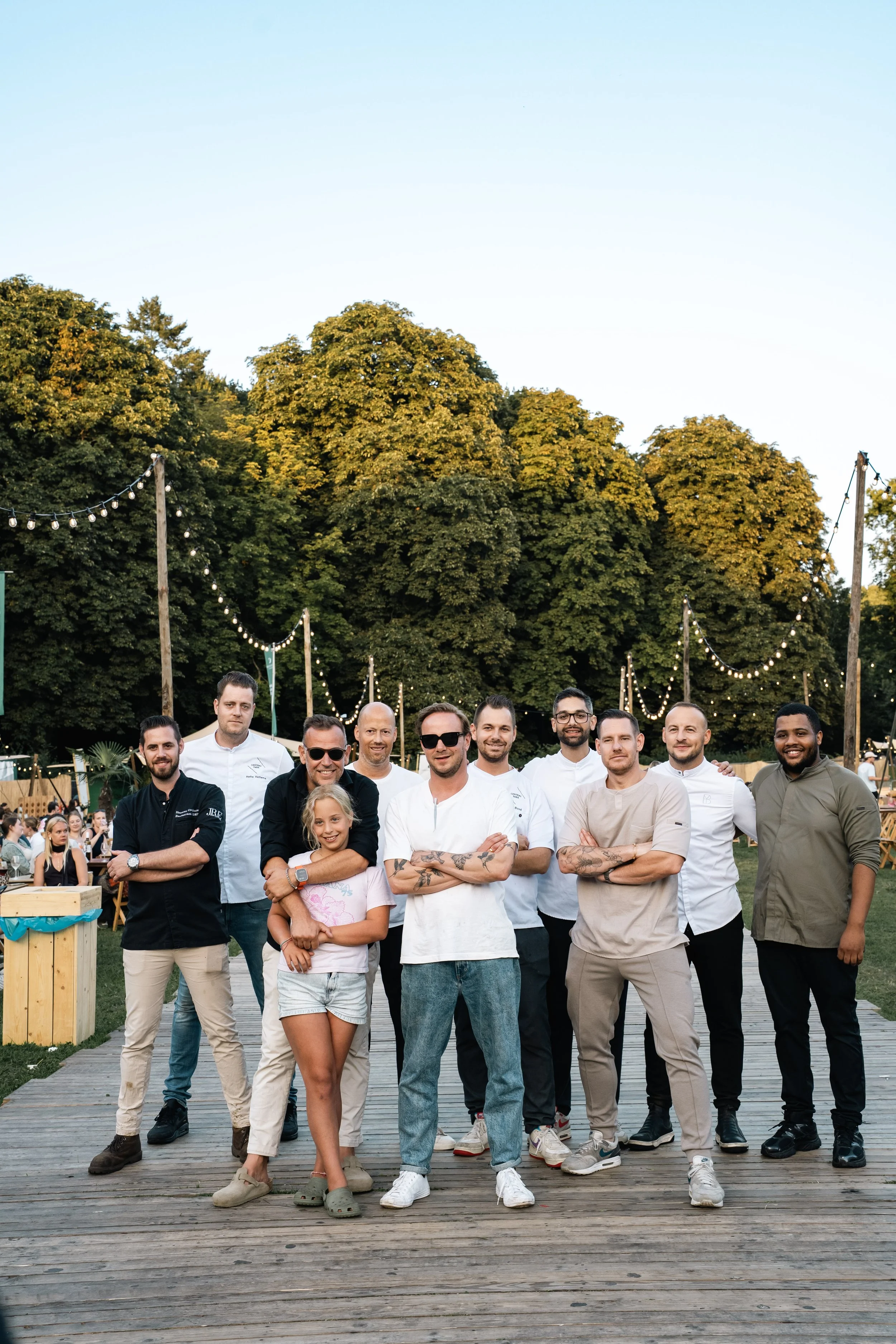 Een groep mensen poseert samen op een houten route in een park met grote bomen en festoonverlichting voor een fotografie of evenementen.