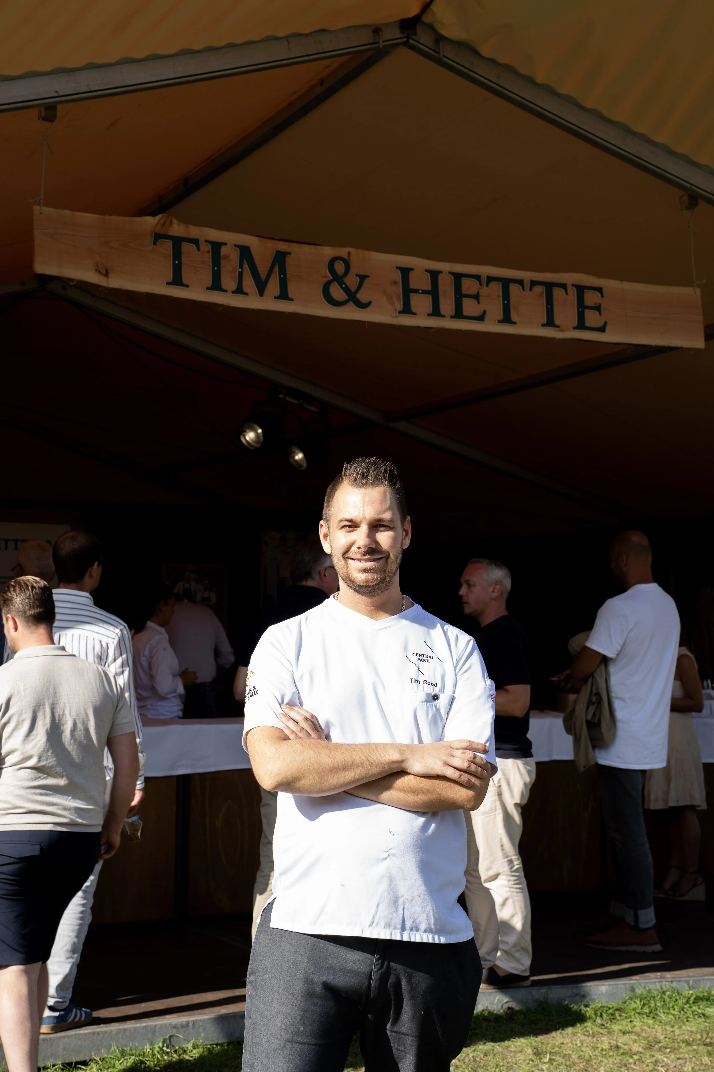 Man in een witte chef-koksjas met armen gekruist, staand buiten voor een houten bar met een groep mensen die eromheen staan, onder een gele luifel met een houten sign met de tekst 'Tim & Hette'.