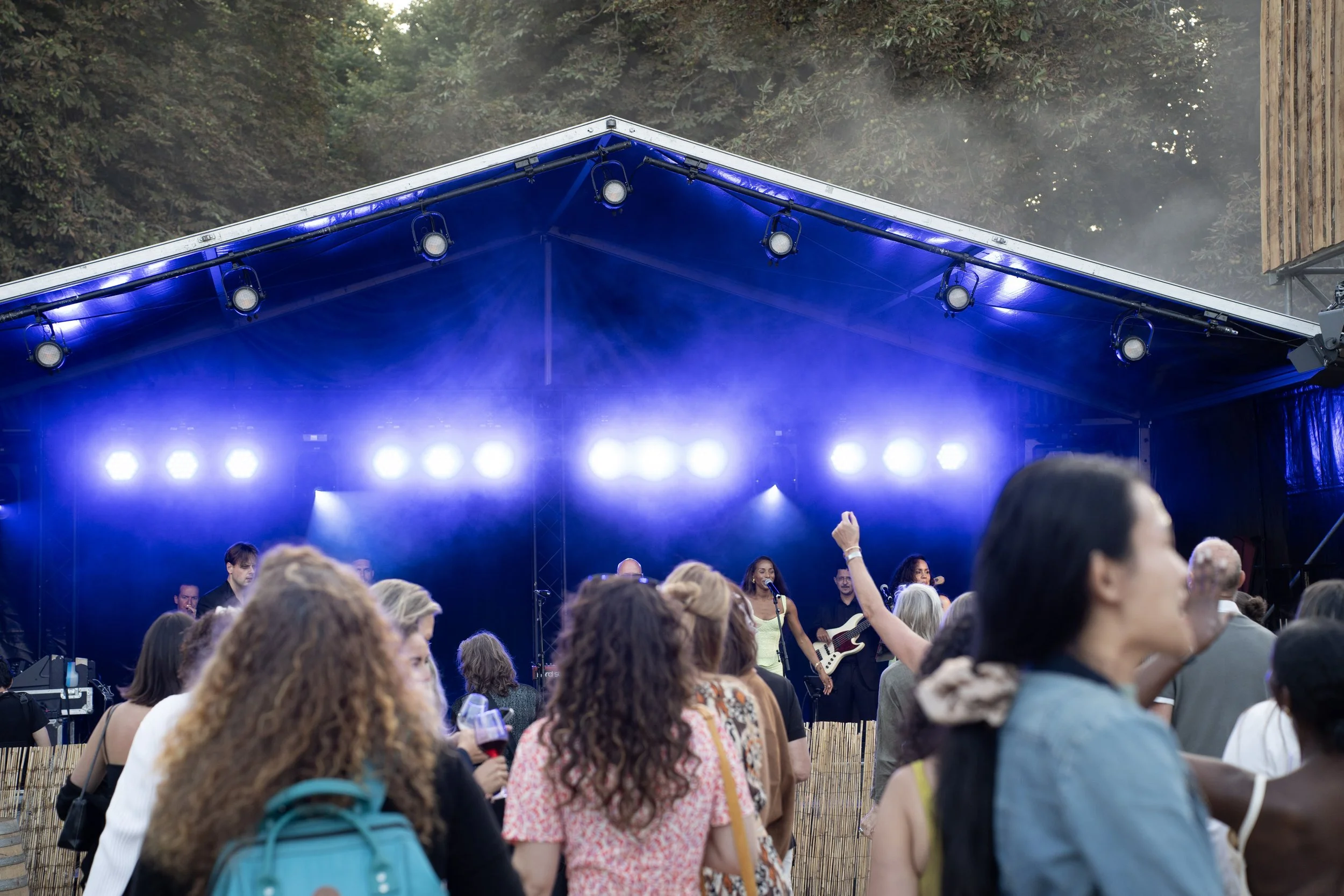 Een buitenpodium met blauwe overkapping en veel mensen in het publiek. Op het podium spelen muzikanten met gitaren en zingen.